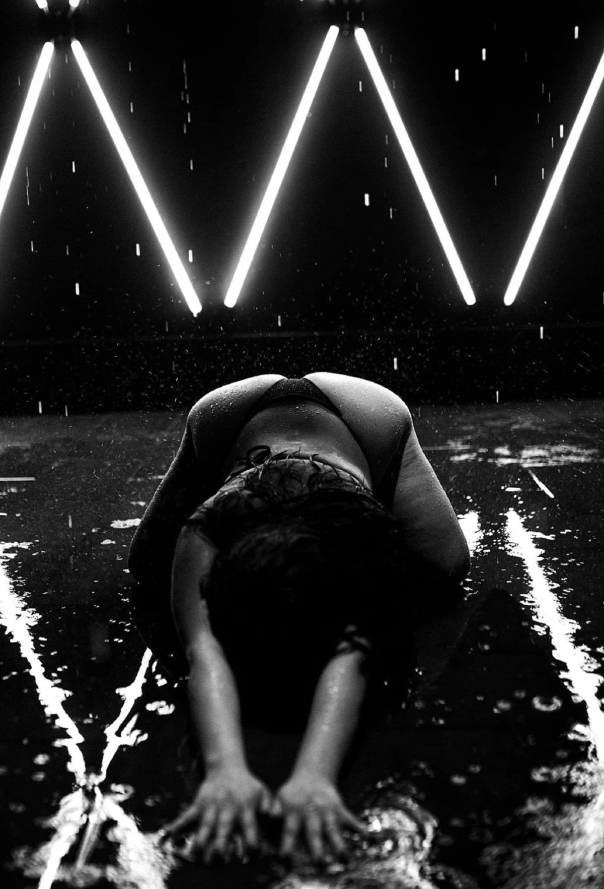 woman in black thong bottoms poses on hands and knees on wet concrete with neon lights during rain room boudoir at Radical Babe Studios