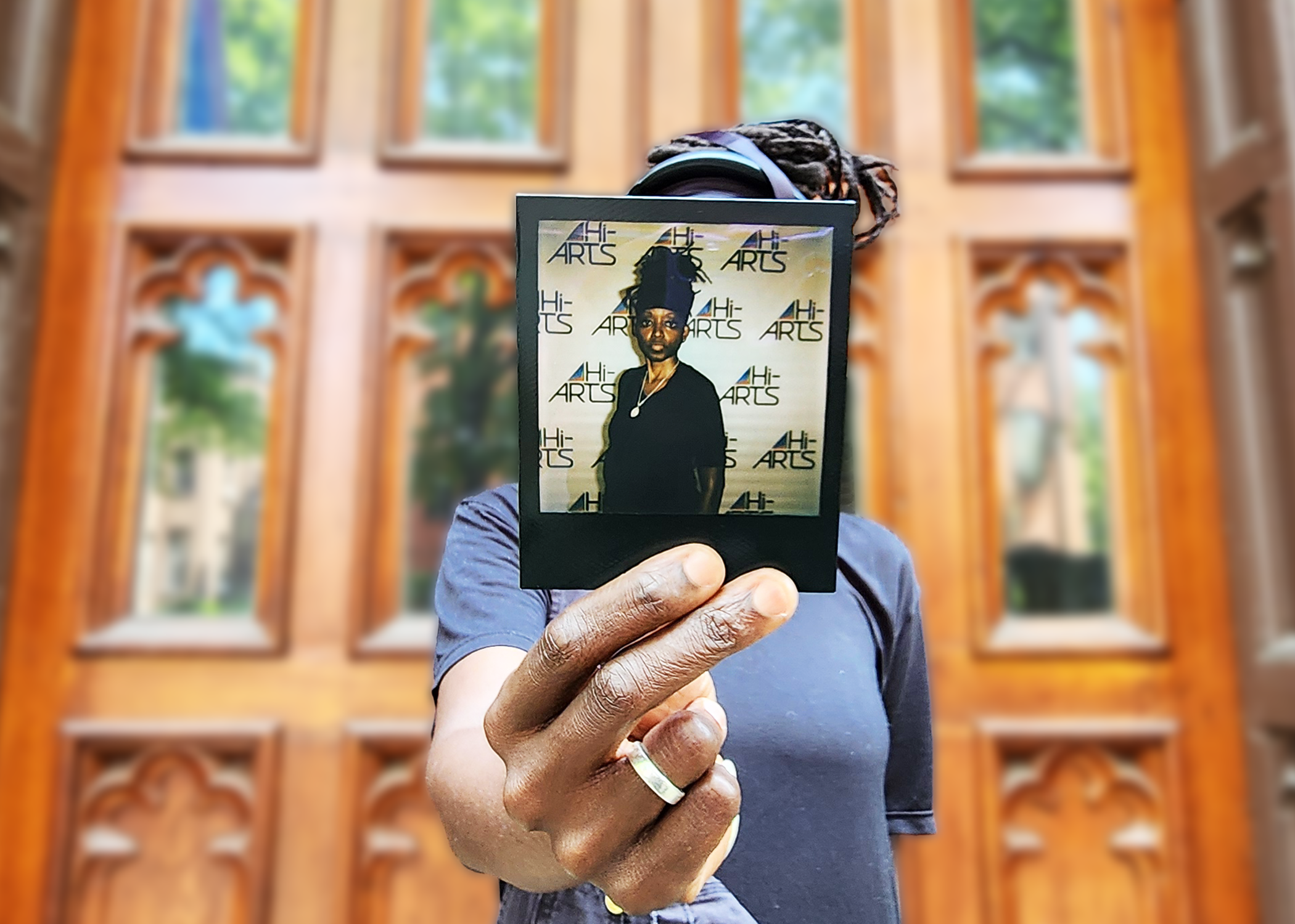 A photo of Debbie holding a Polaroid of herself standing in front of her face. She is wearing black.