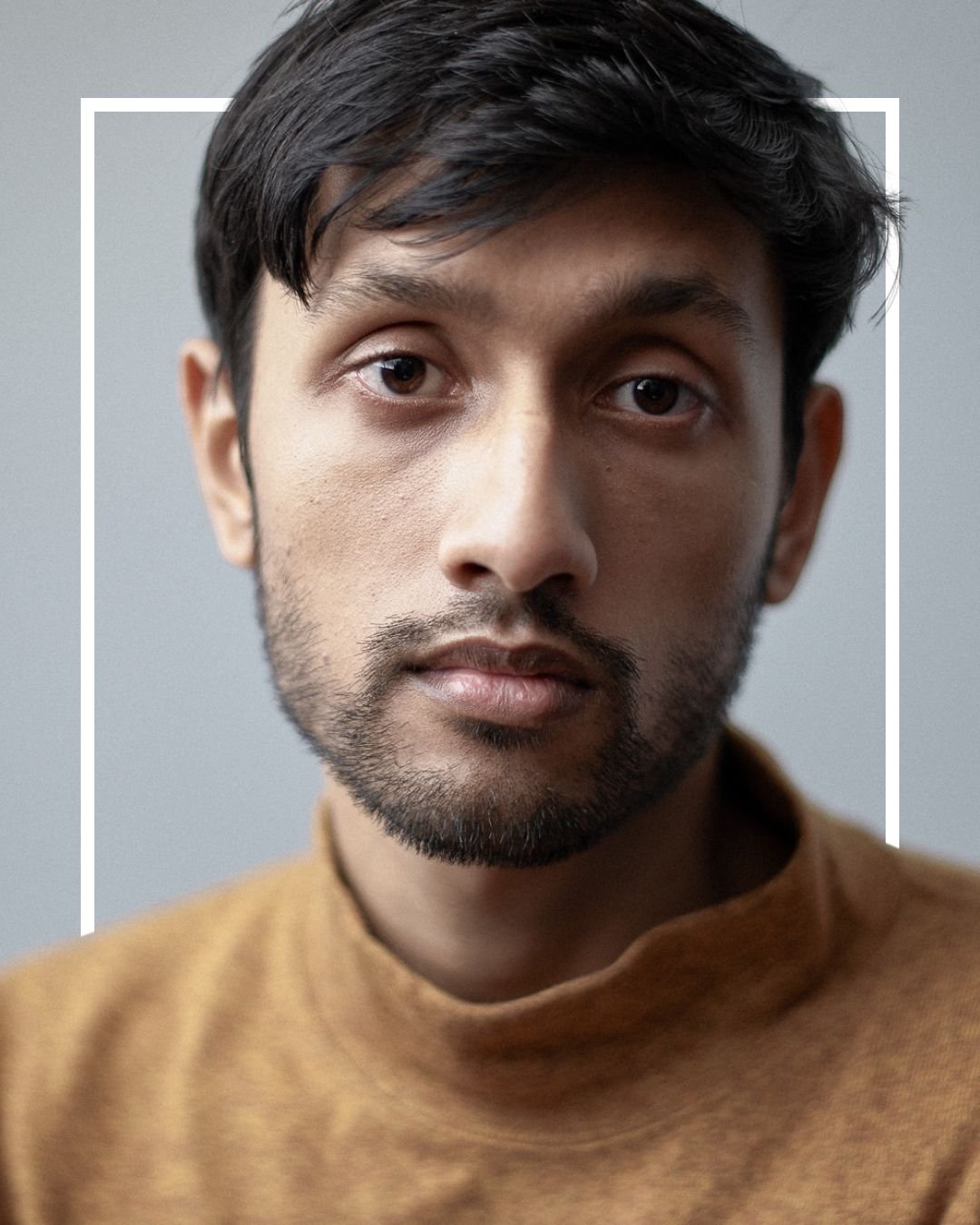 A close up color photograph of Nikhil in front of a grey backdrop. They have short black hair and a short beard. They are wearing a tan turtleneck.