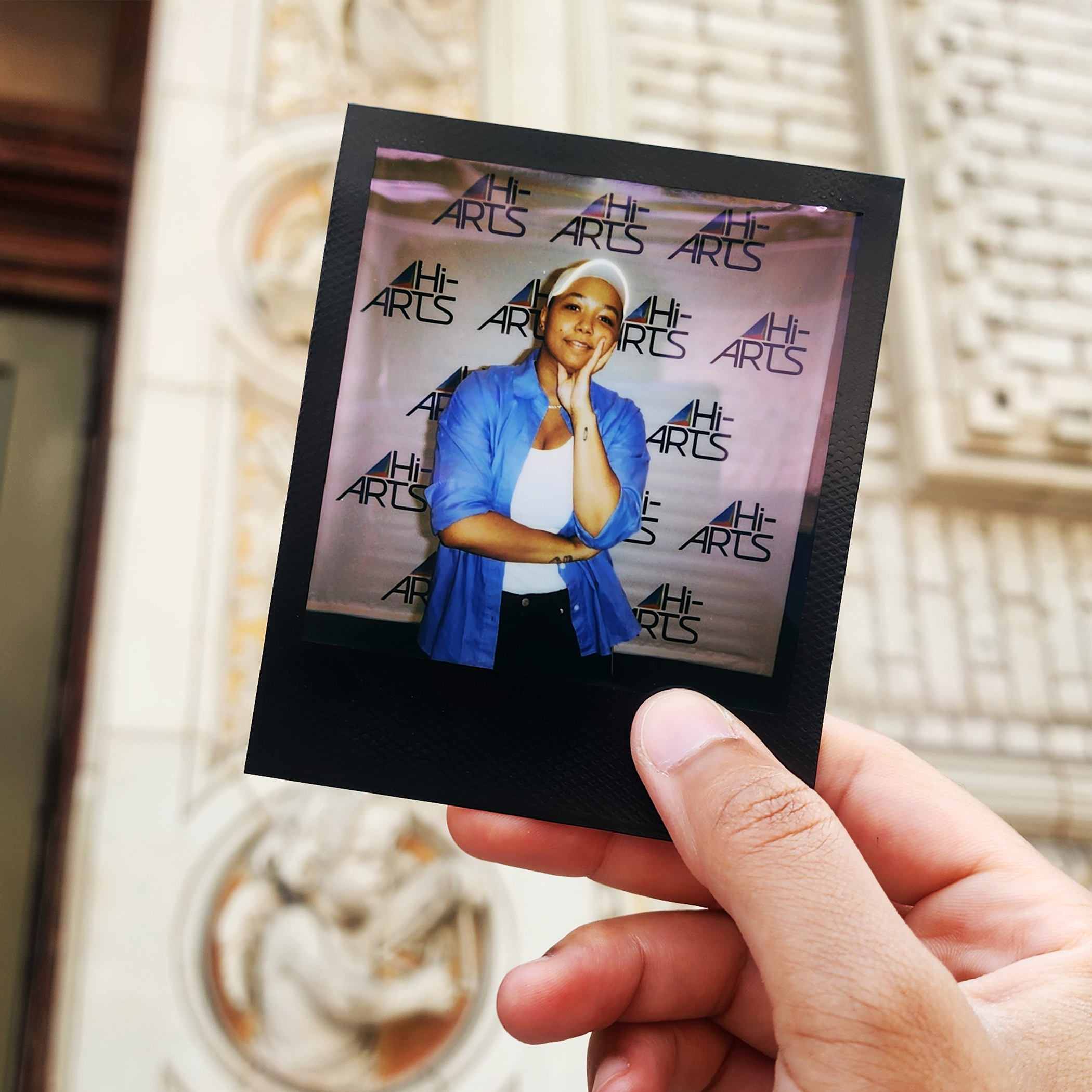 A photo of Robyn's hand holding a Polaroid of a photo of her in front of a brick facade. She is wearing a blue shirt and a white cap.