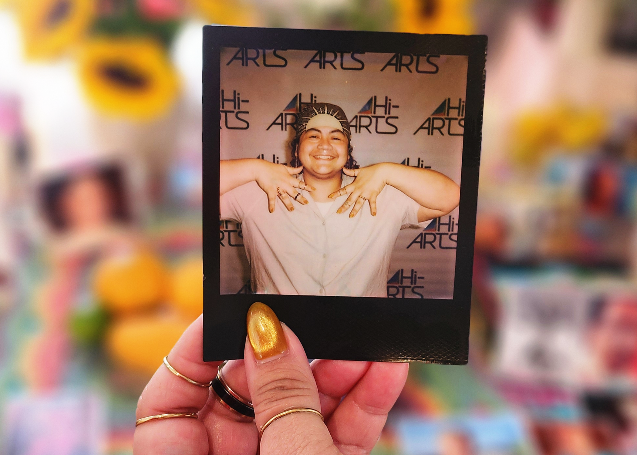 A photo of MJ's hand holding a Polaroid of themselves. They are wearing a white shirt and a brown and golden headscarf.