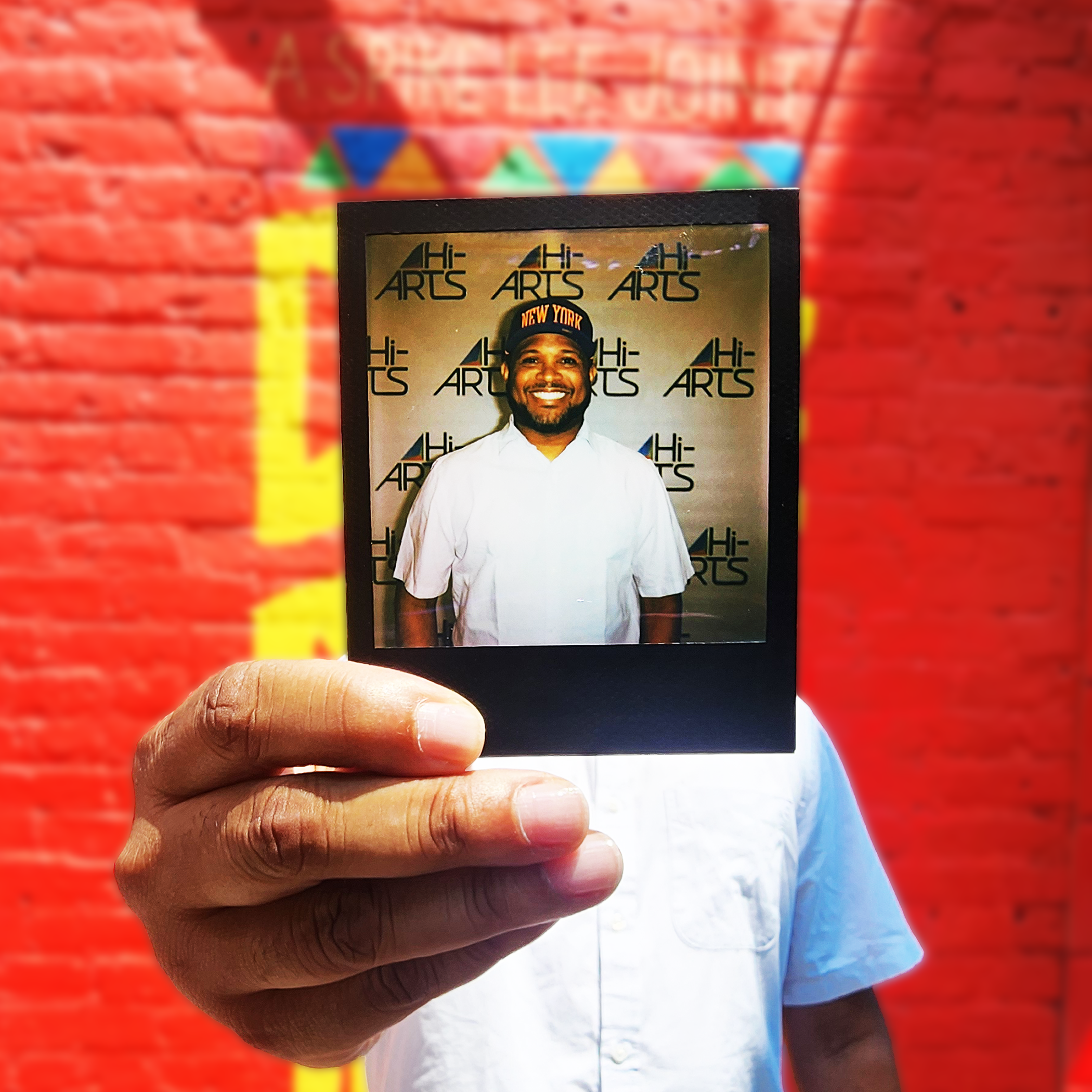 A photo of Aaron holding a Polaroid of himself in front of his face. He is wearing a white tshirt and is standing in front of a bright red wall.