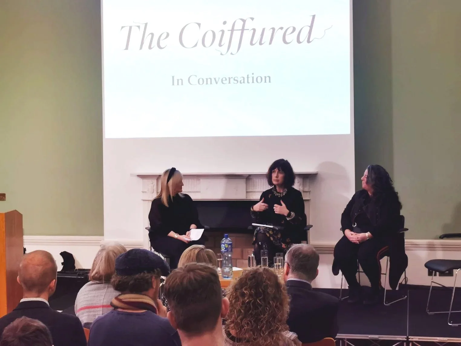 Amanda Jane Graham participating in a panel discussion at a conference titled 'The Coiffured: Shifting Perception', with three women seated on stage in front of an audience. Amanda Jane Graham is speaking, while the other two women listen.