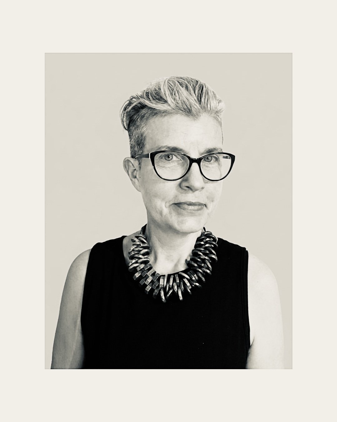 This is a profile black and white photo of Irish artist Amanda Jane Graham. She has short blonde hair and is wearing black framed glasses, black short sleeved top and large chunky necklace.