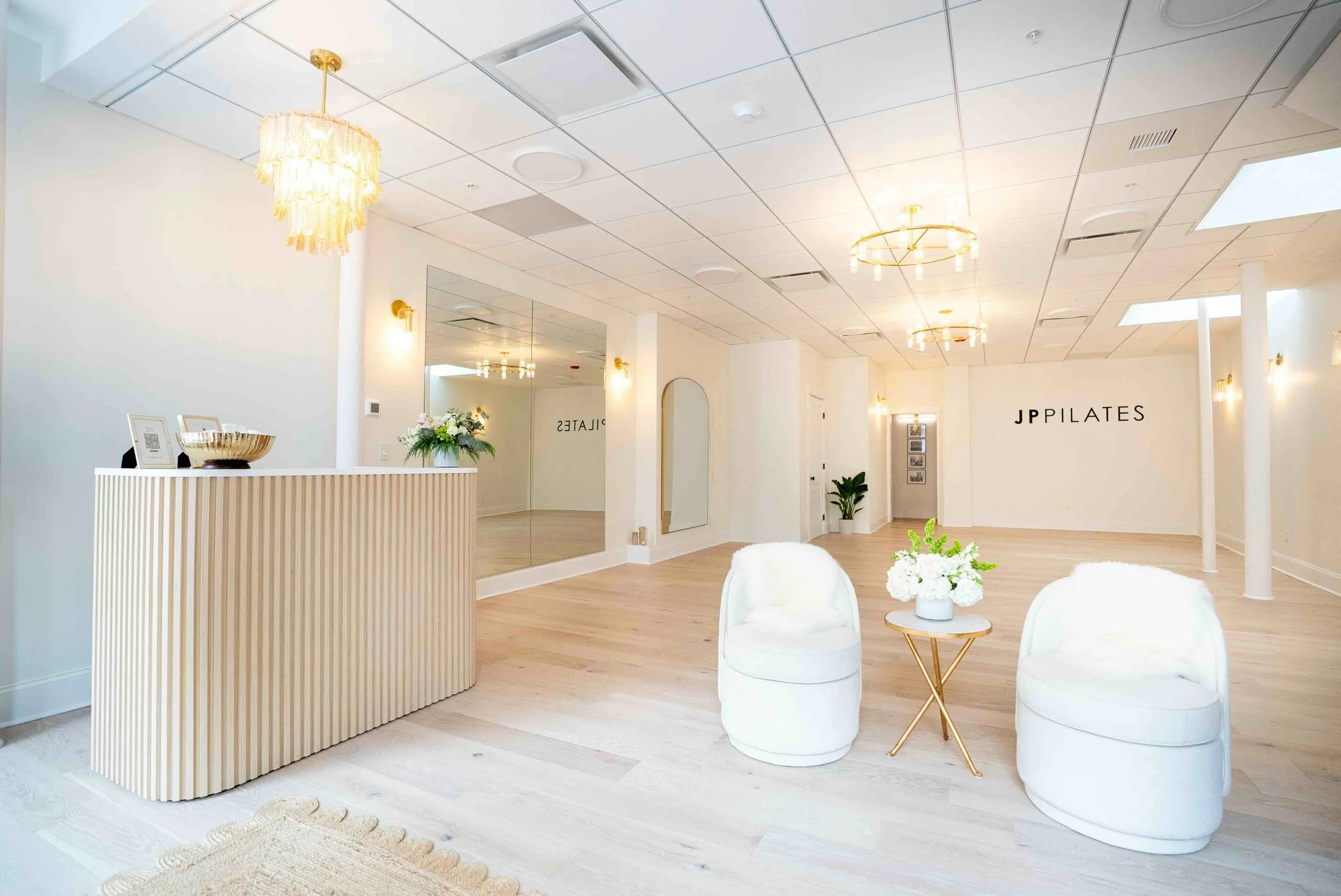 A bright, modern interior space with white walls, light wood flooring, and decorative lighting fixtures. There are white chairs with round tables topped with white flowers, a cream-colored rug, and a reception desk with a bowl and framed QR code. The background features a large mirror and a wall with the text 'JP PILATES'.