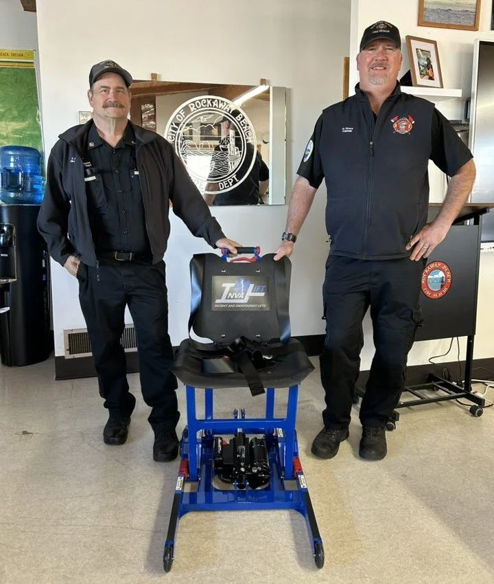 Two firefighters standing indoors, smiling, with a rescue device on a chair and a small blue stretcher or spine board. Behind them are water bottles, a desk, and framed pictures on the wall, including a city emblem for Rockaway Beach.
