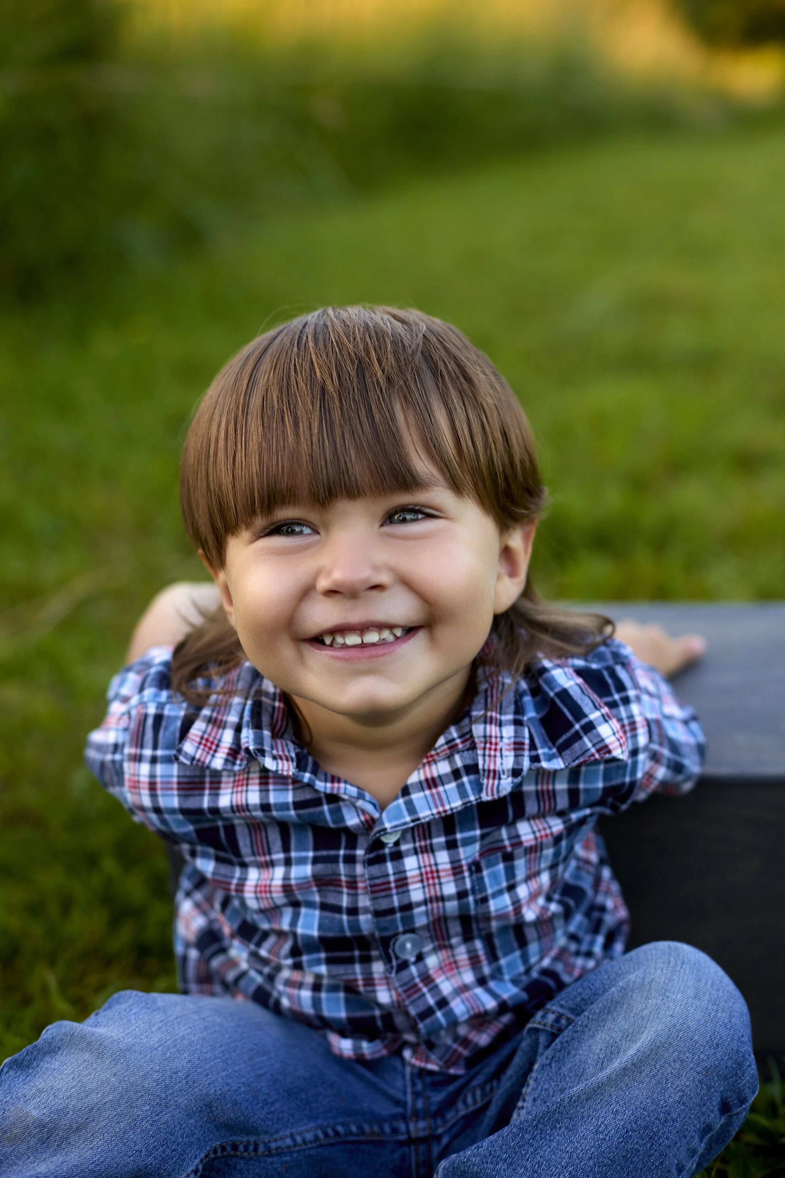 A young boy with brown hair, wearing a plaid shirt and blue jeans, smiling and lying on grass with his head slightly tilted upward and hands behind him, in an outdoor setting with blurred greenery in the background. Ada Hayden Park in Ames, Iowa.