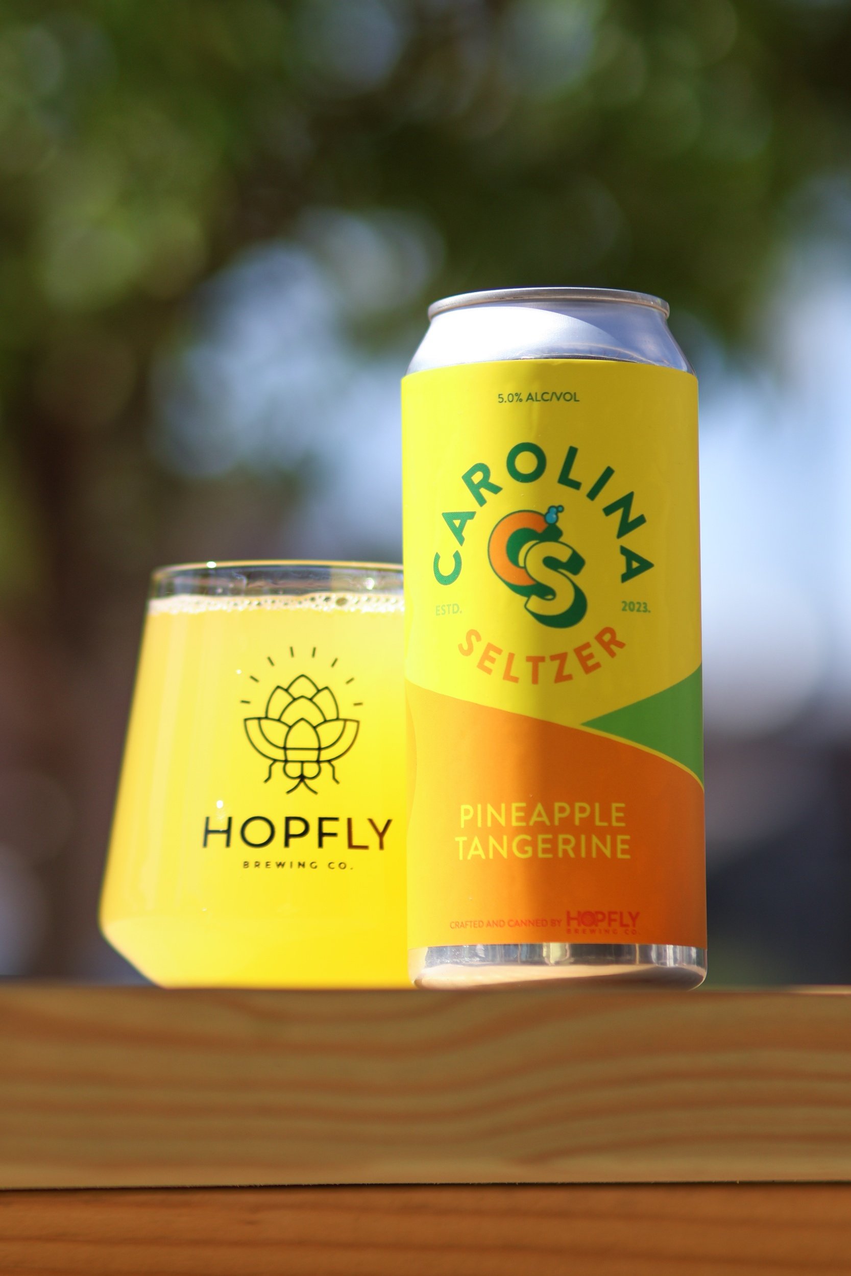 HopFly Brewing Co. Carolina Seltzer in a pineapple tangerine flavor can next to a glass with the brewery's logo.
