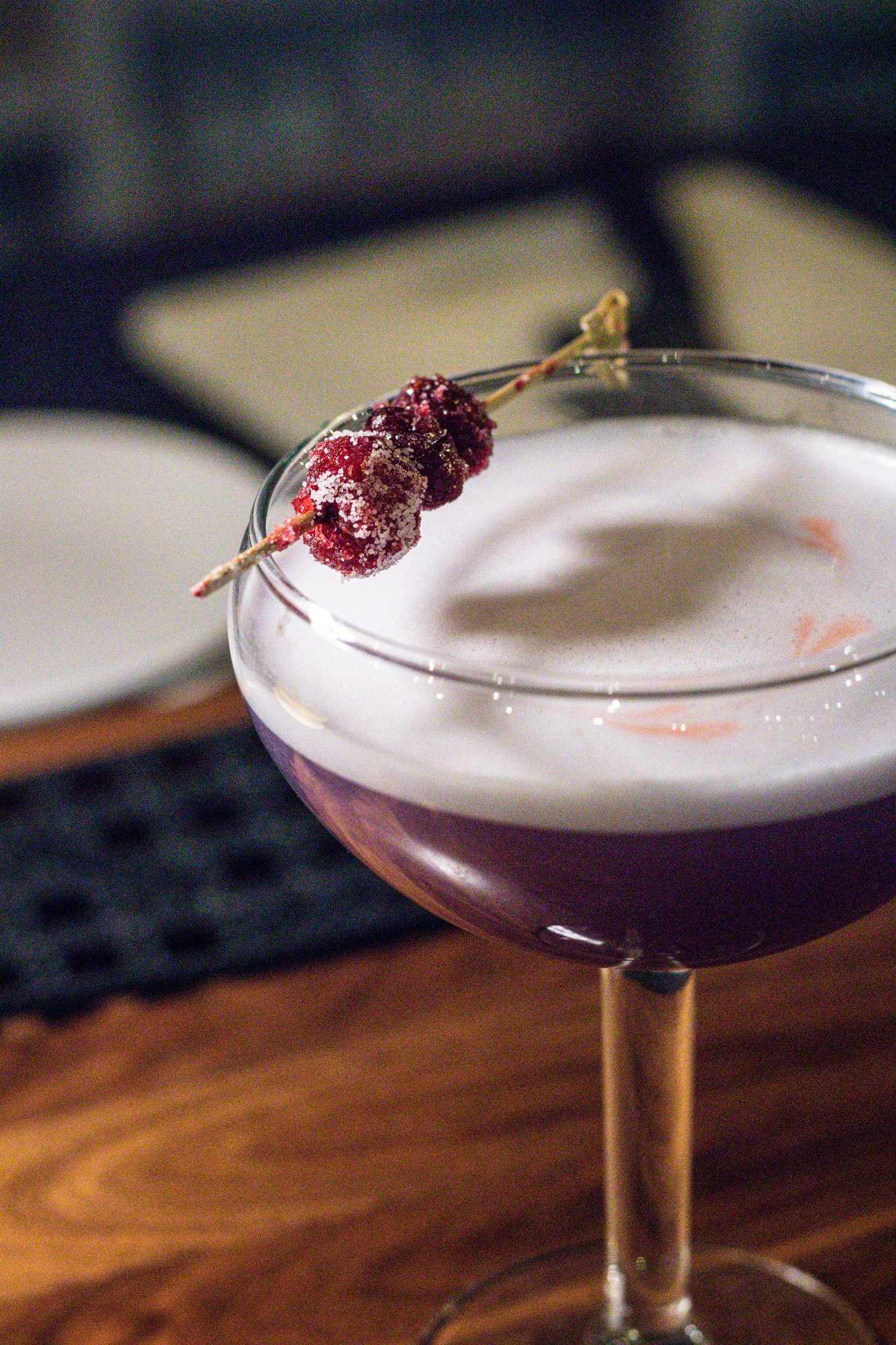 Purple cocktail in a coupe glass with a frothy top, garnished with sugared cranberries on a skewer.