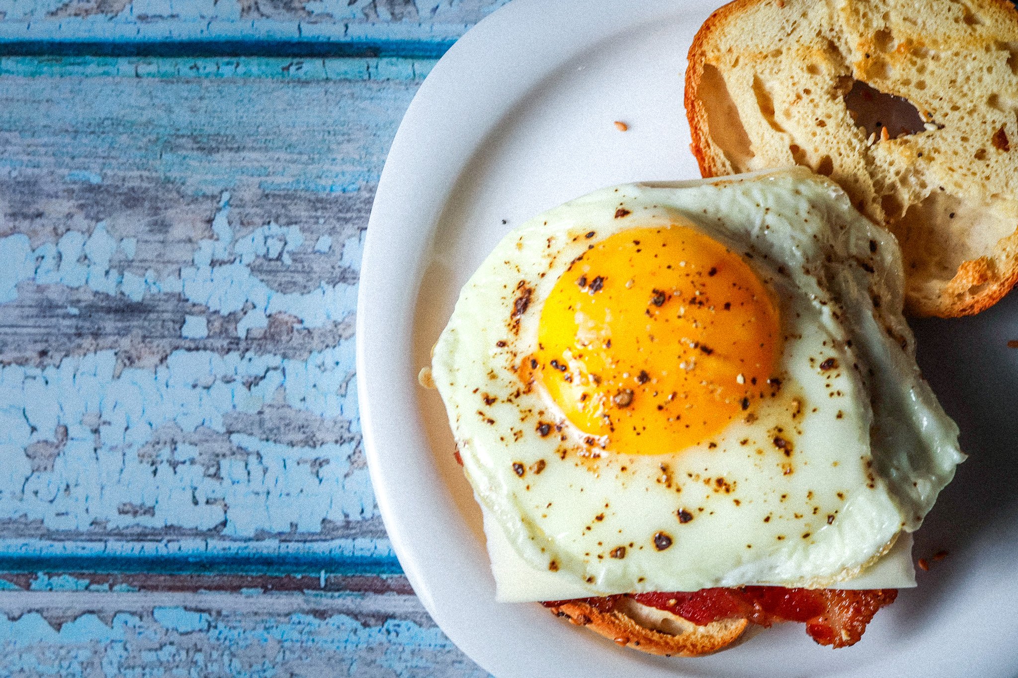 Fried egg on a bagel sandwich with cheese and bacon on a white plate, blue weathered wood background.