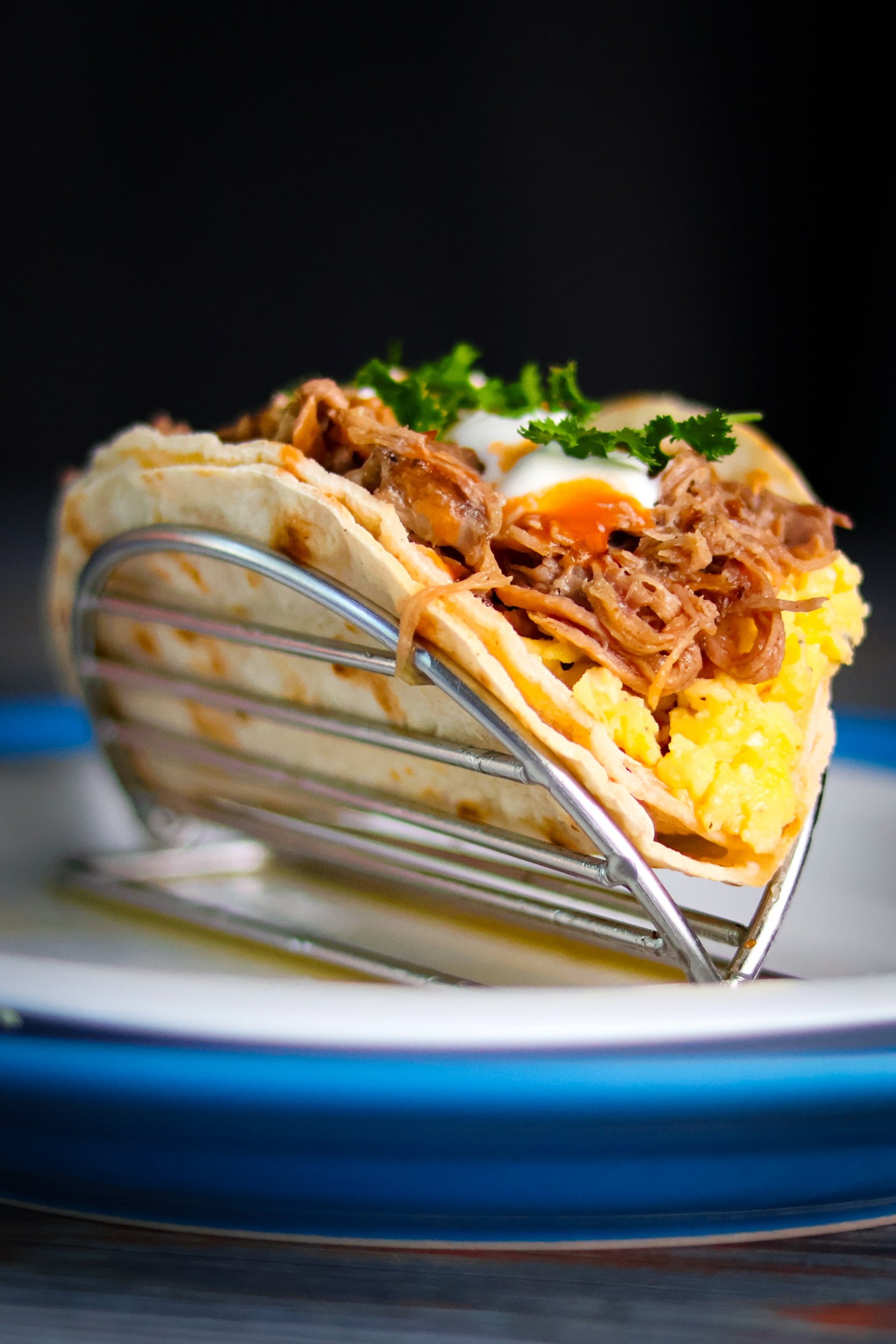 A taco with shredded meat, scrambled eggs, and parsley in a metal holder on a plate.