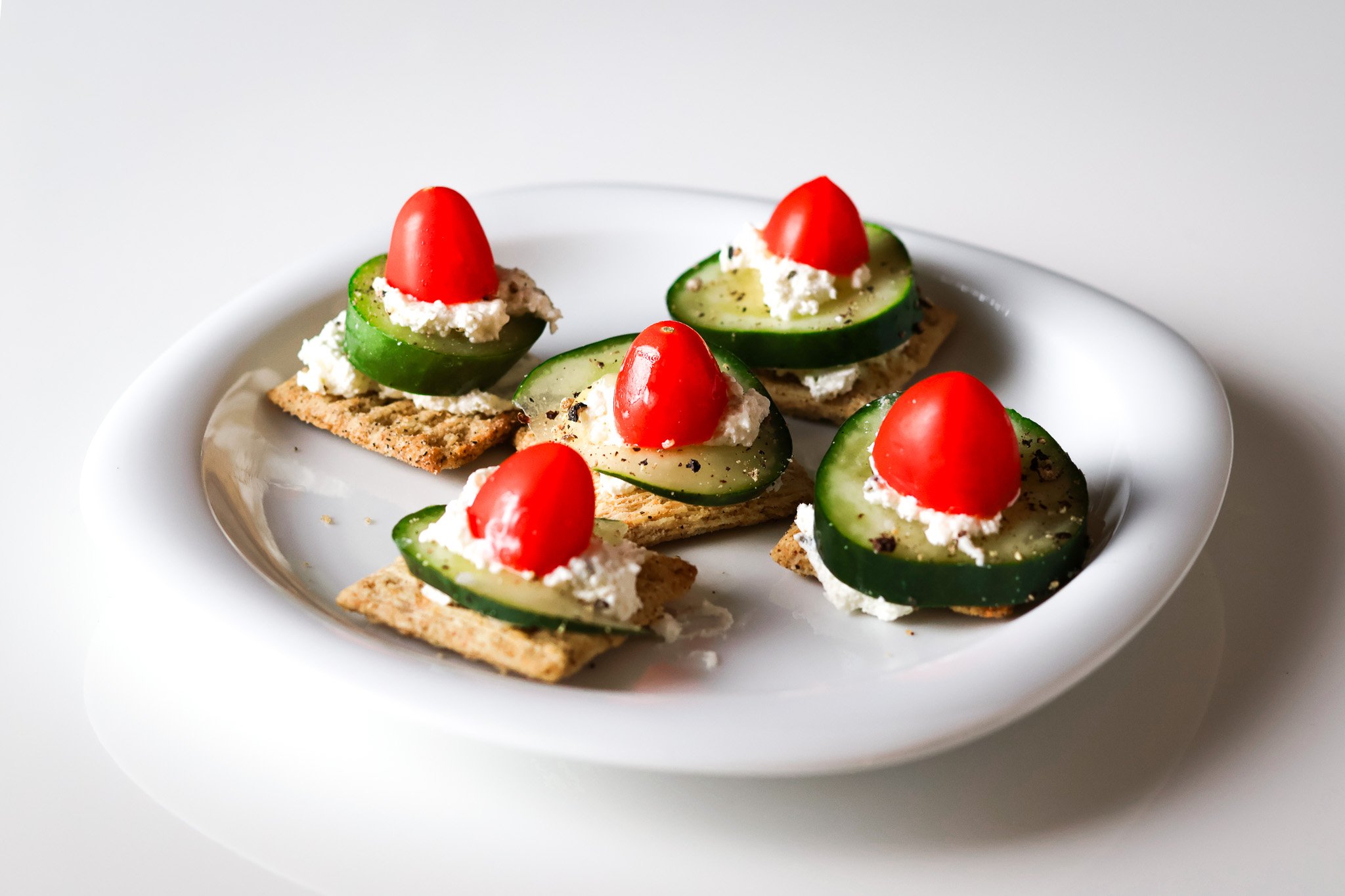 Plate of crackers topped with cucumber slices, cheese, and cherry tomato halves.