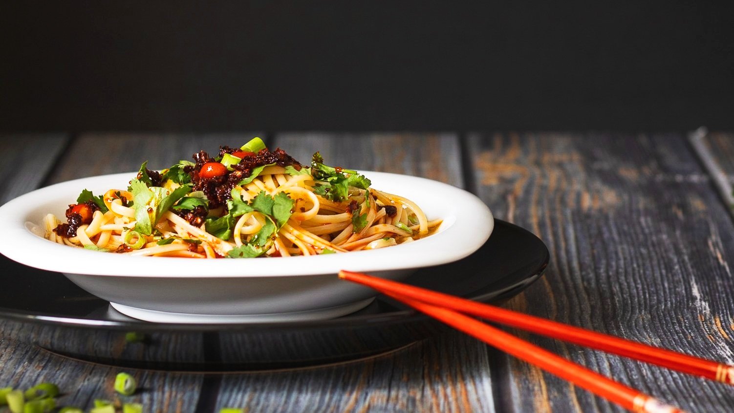 A plate of Asian-style noodles garnished with fresh cilantro, chopped green onions, and red chili peppers, with a pair of red chopsticks placed nearby.