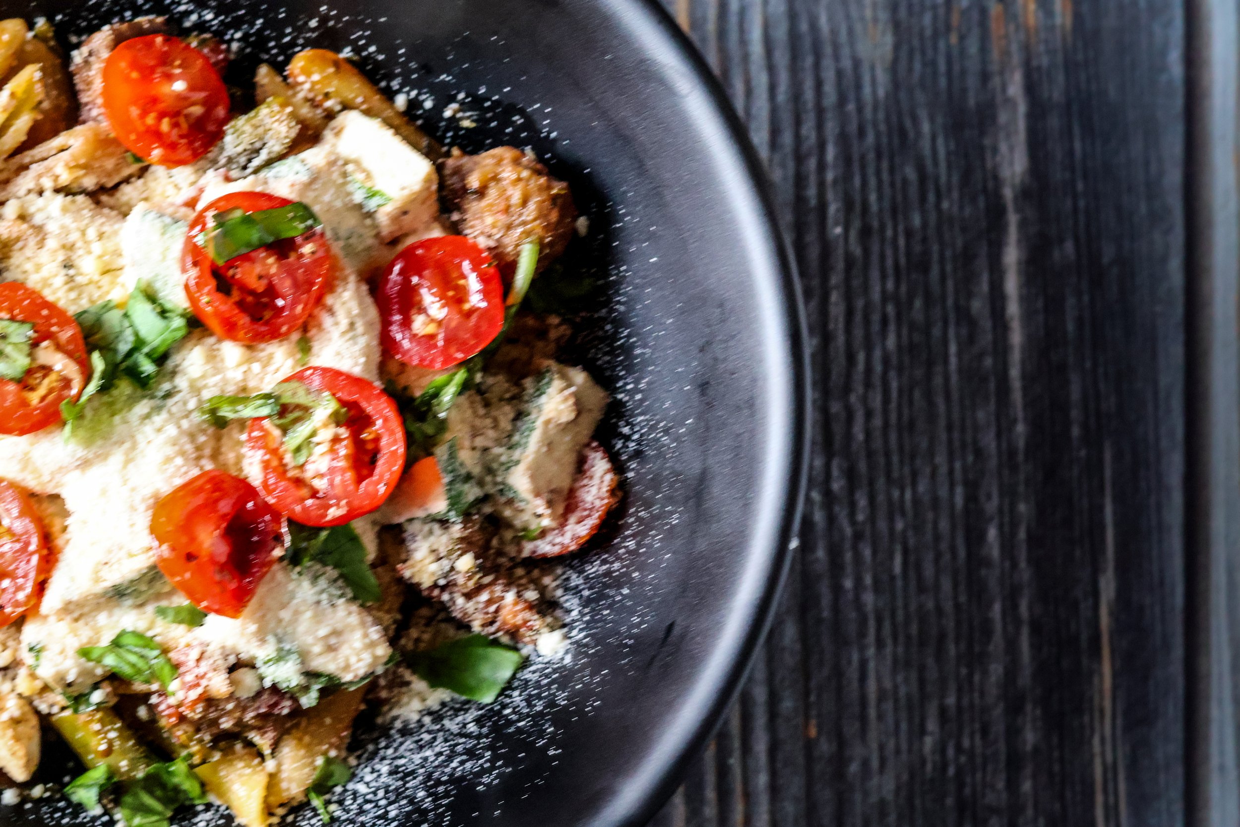 Close-up of a bowl of pasta with cherry tomatoes, herbs, and grated cheese on a dark textured surface.
