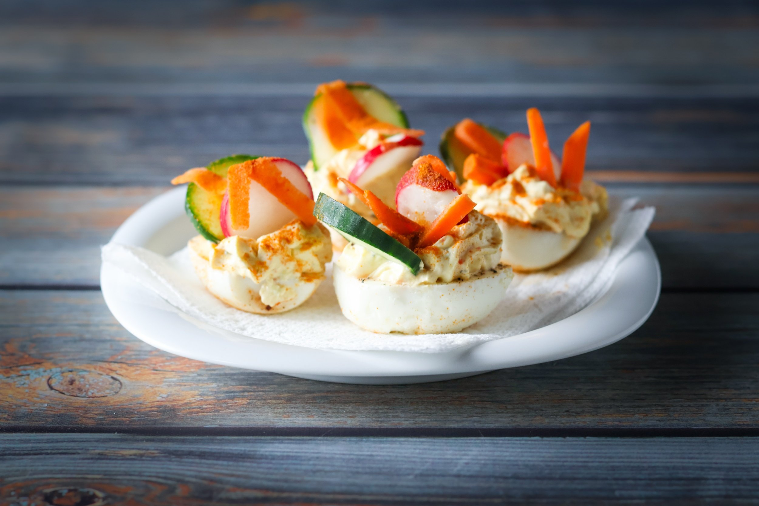 Deviled eggs topped with sliced radishes, cucumbers, and carrots on a white plate.