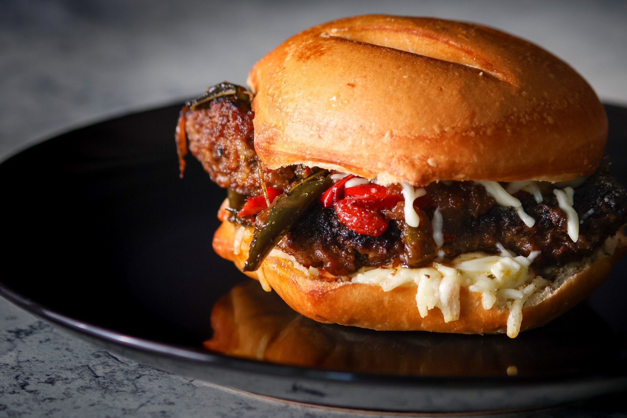 Close-up of a spicy Italian sausage sandwich with peppers and mozzarella cheese on a bun, served on a black plate.