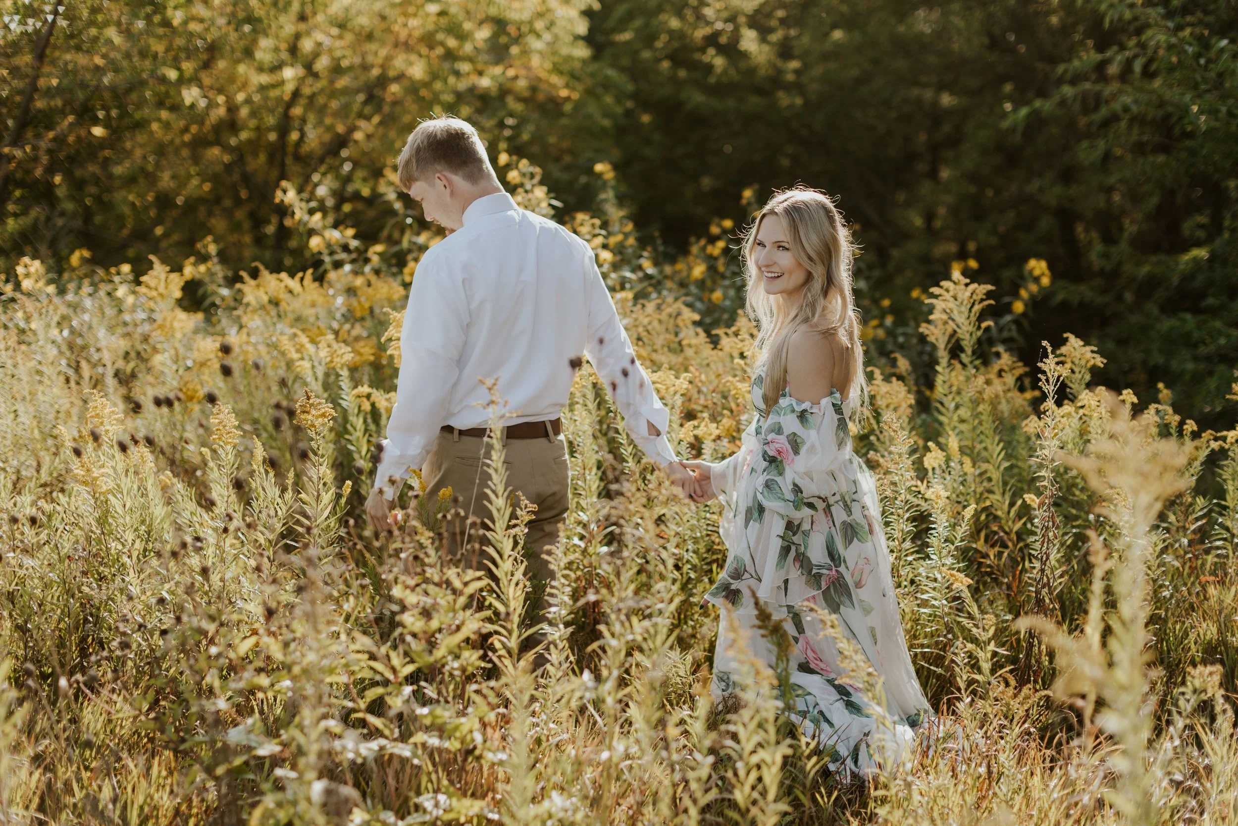 young couple in bridal attire holding hands and walking through field at golden hour