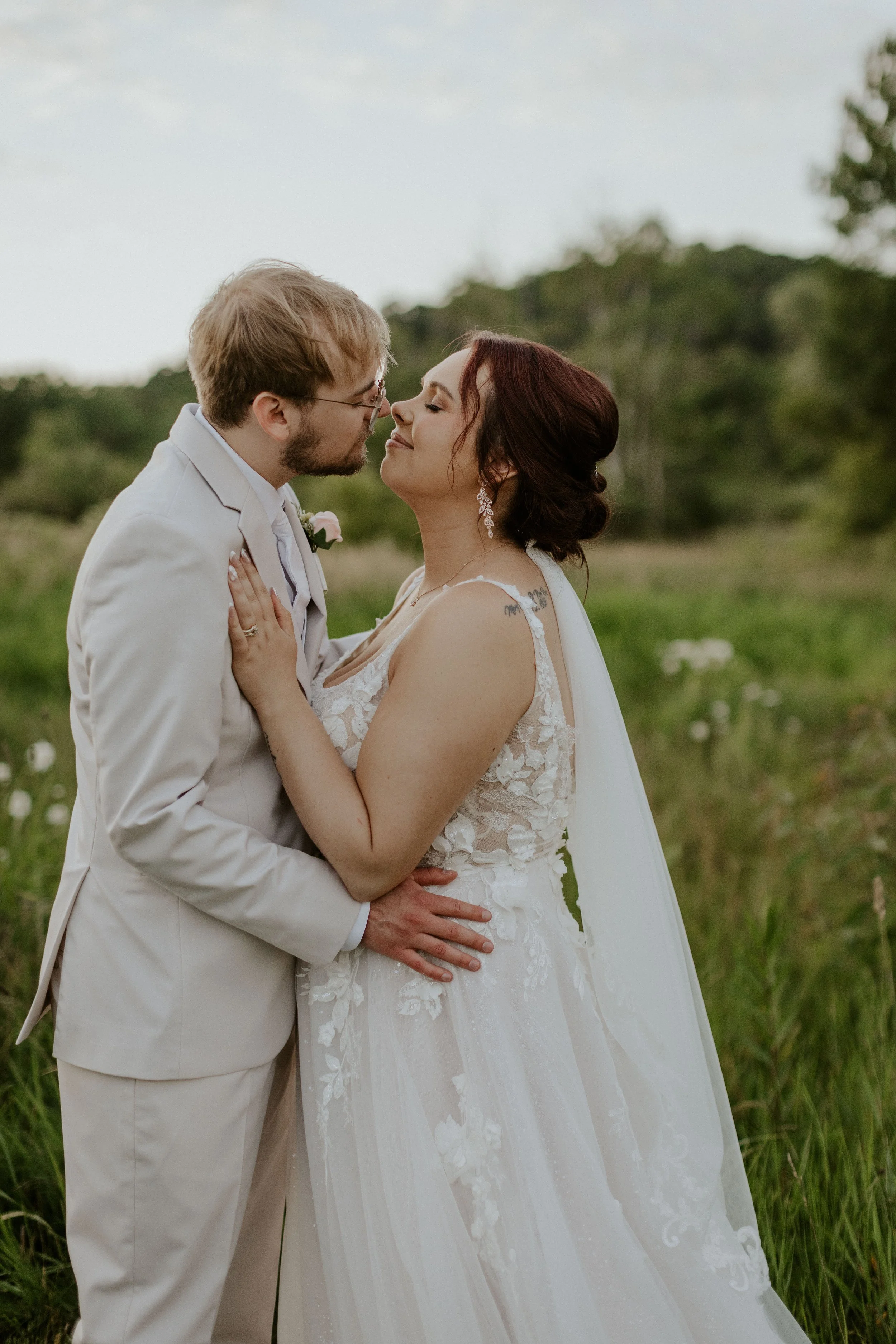 young couple in bridal attire embracing and leaning in for kiss in a grassy meadow