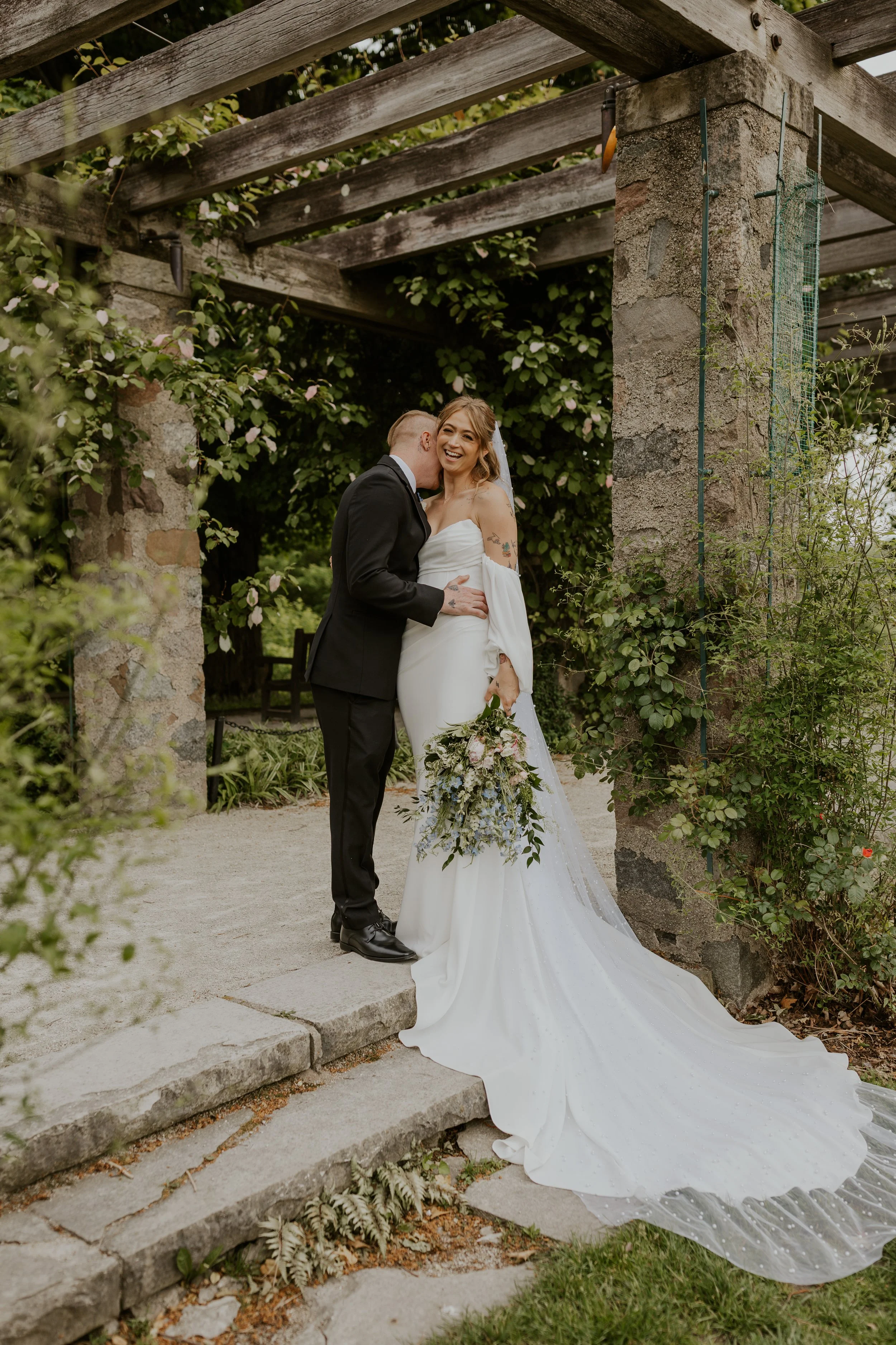 couple dressed in bridal attire laughing underneath pergola covered in ivy