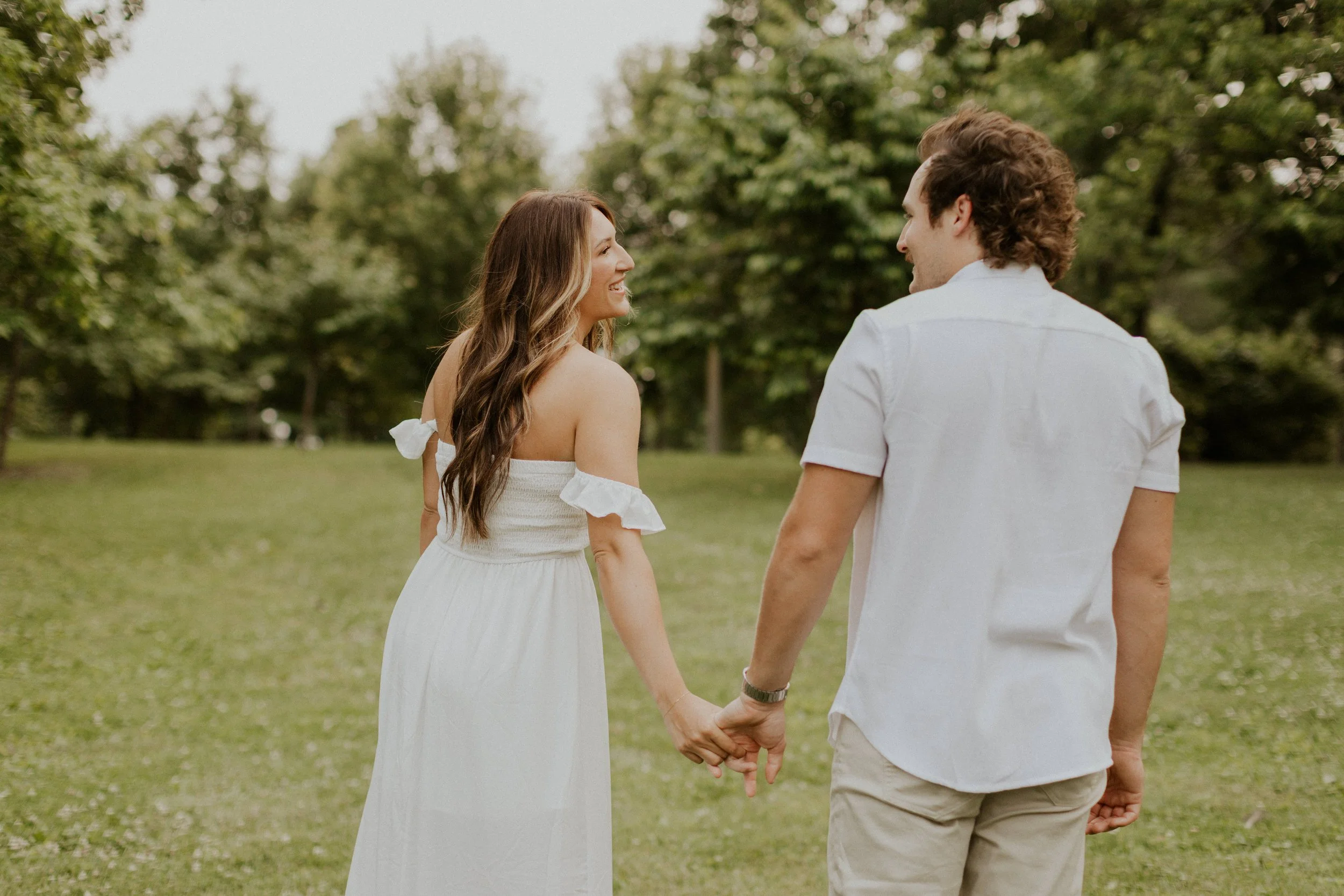 young couple dressed in white holding hands and laughing while walking in park in Fort Collins
