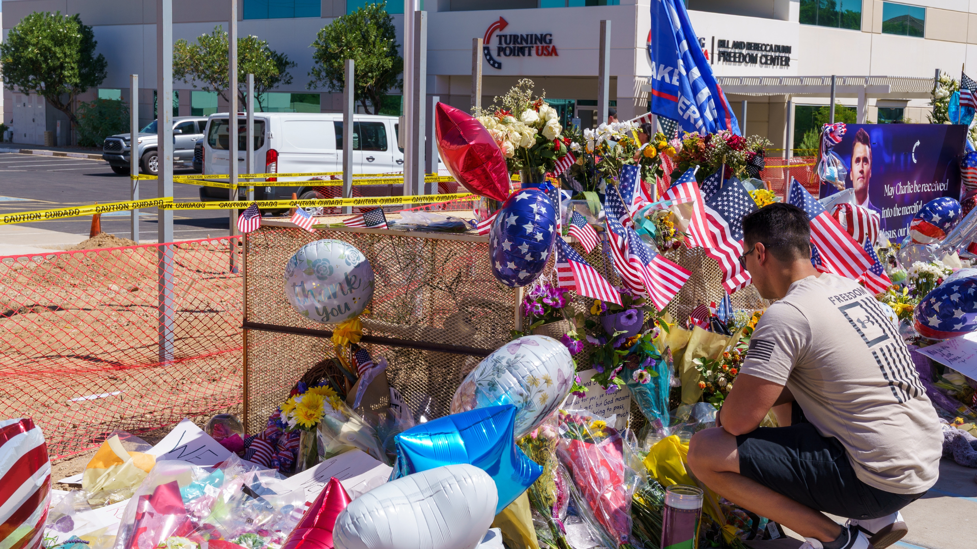 young man kneeling among flowers and momentos left at a memorial to charlie kirk