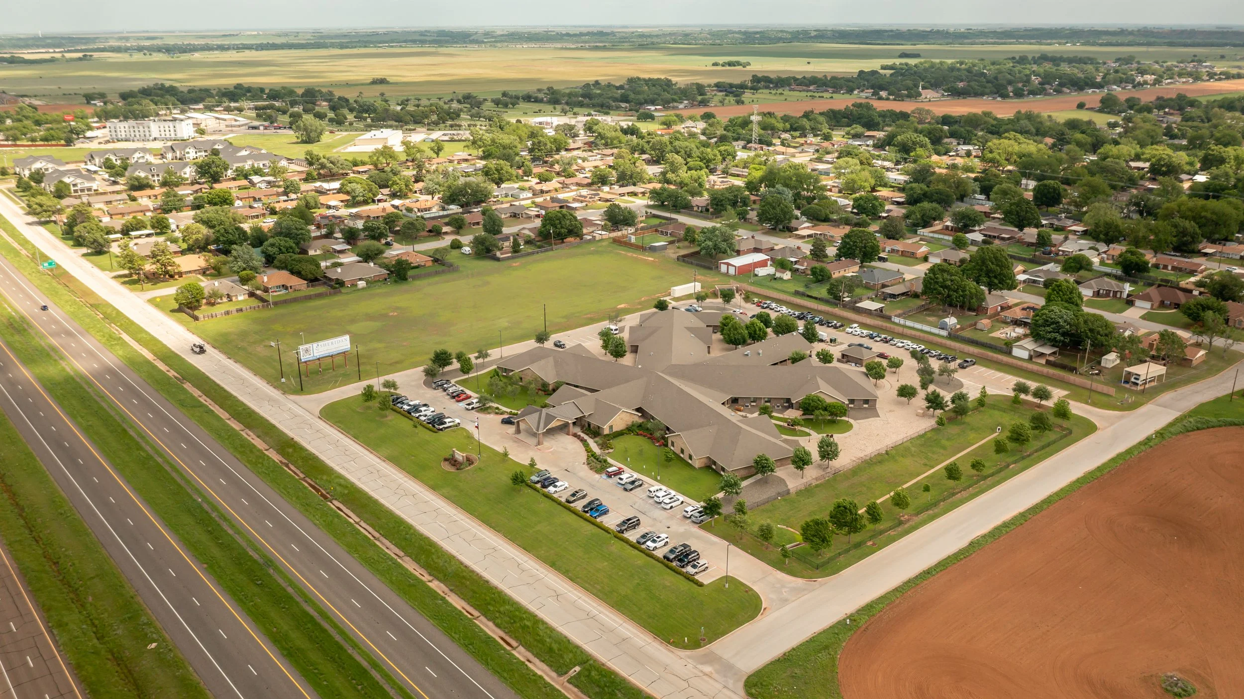 Aerial view of a suburban residential area with houses, green trees, and a large building, next to a highway and open farmland in the background.