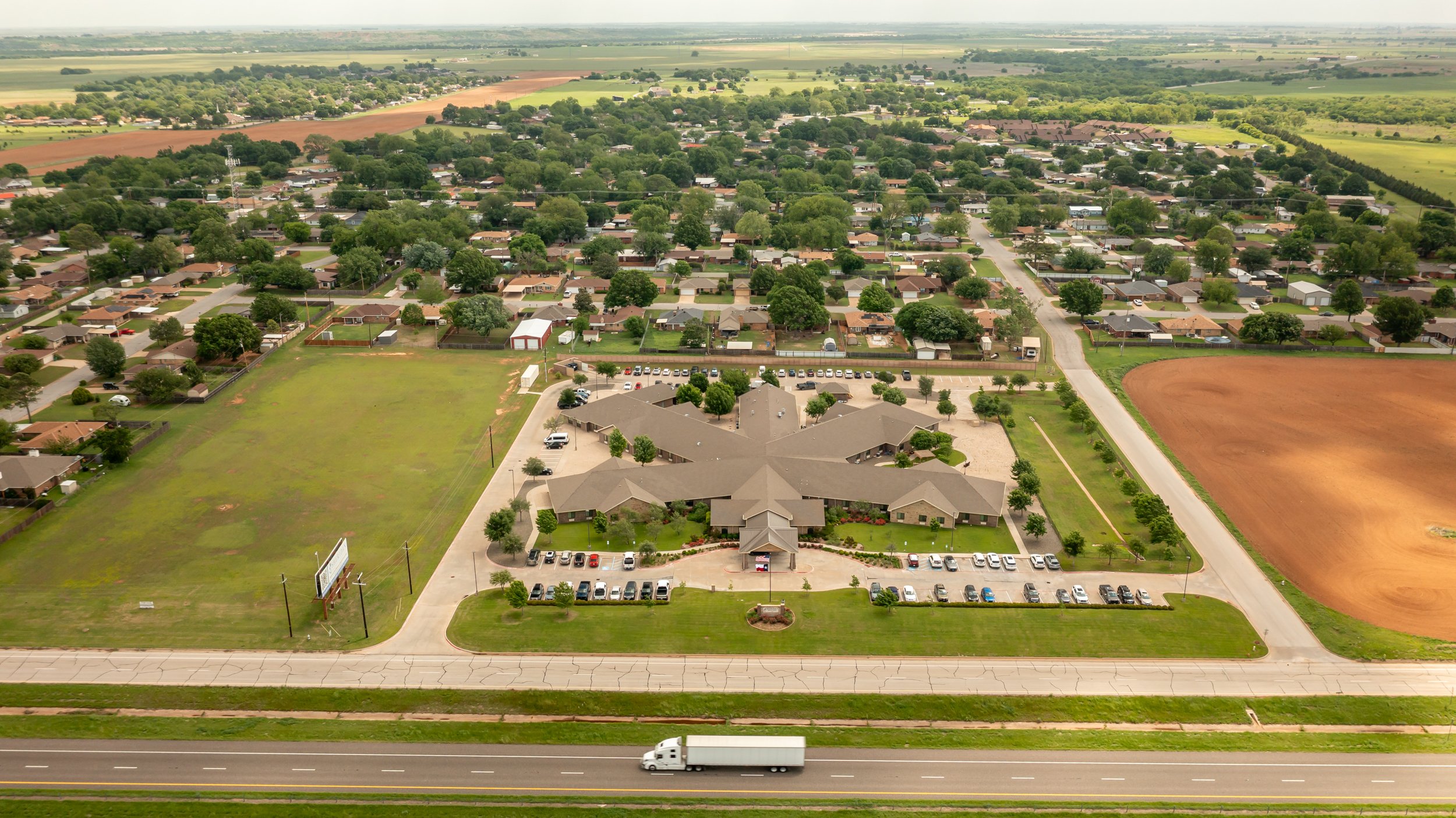 An aerial view of a suburban neighborhood with houses, green trees, parking lots, and open fields, with a busy highway at the bottom.