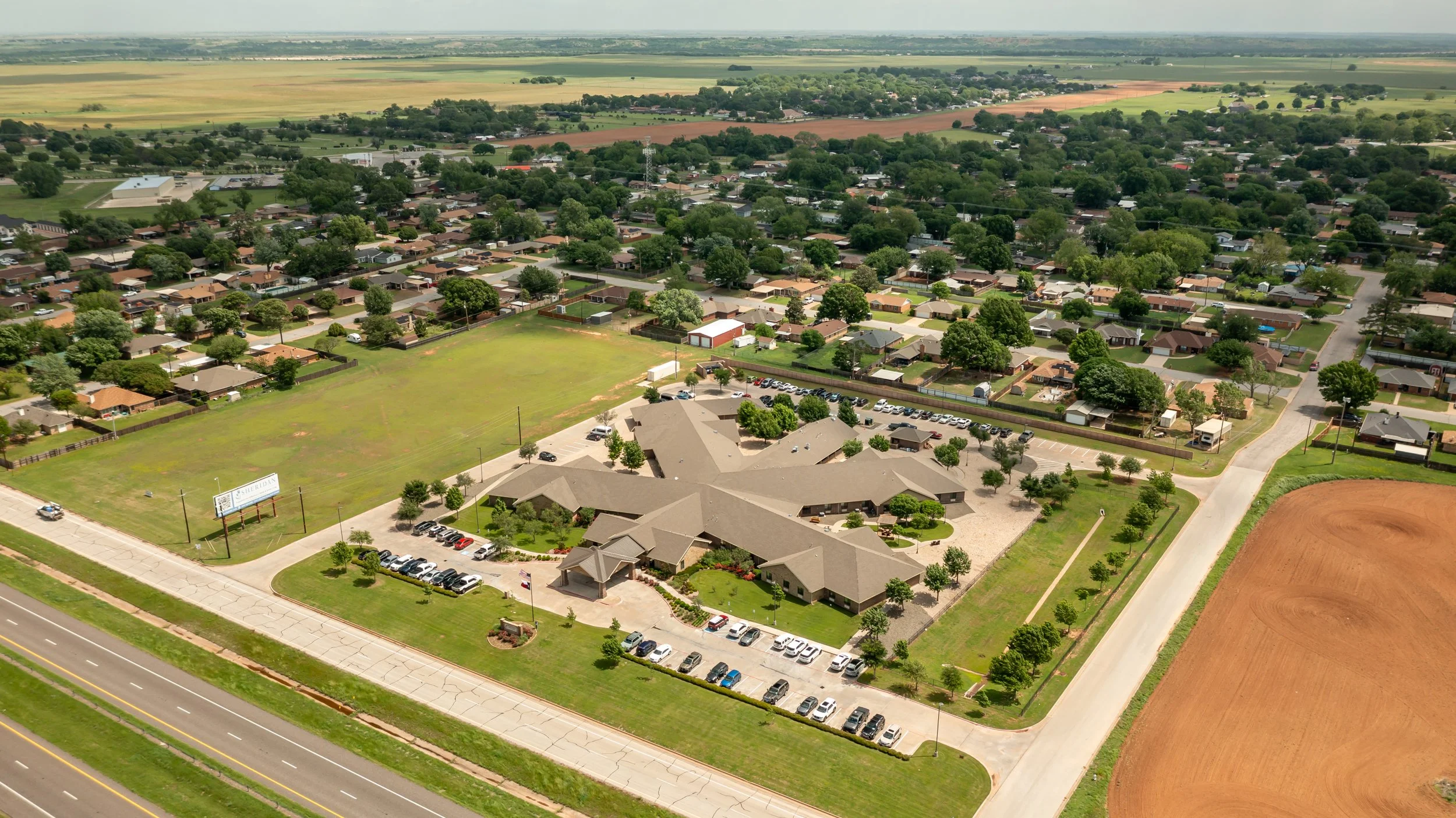 Aerial view of a suburban neighborhood with residential houses, parking lots, green trees, and open fields, along a highway.