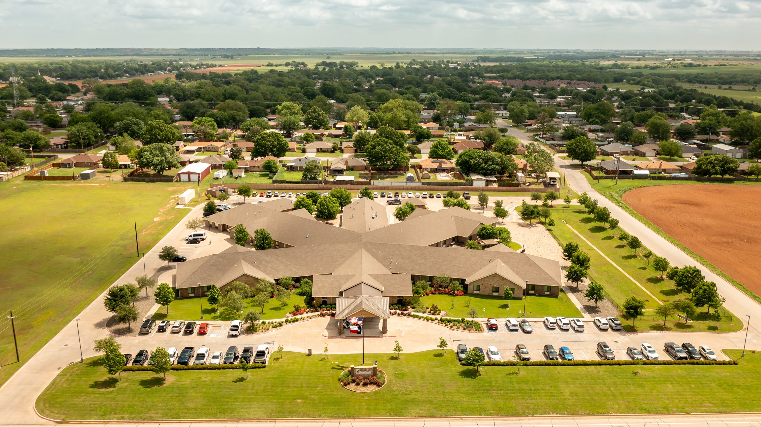 Aerial view of a suburban neighborhood with a central building, surrounded by parking lots, green lawns, and homes, with fields and trees in the background.