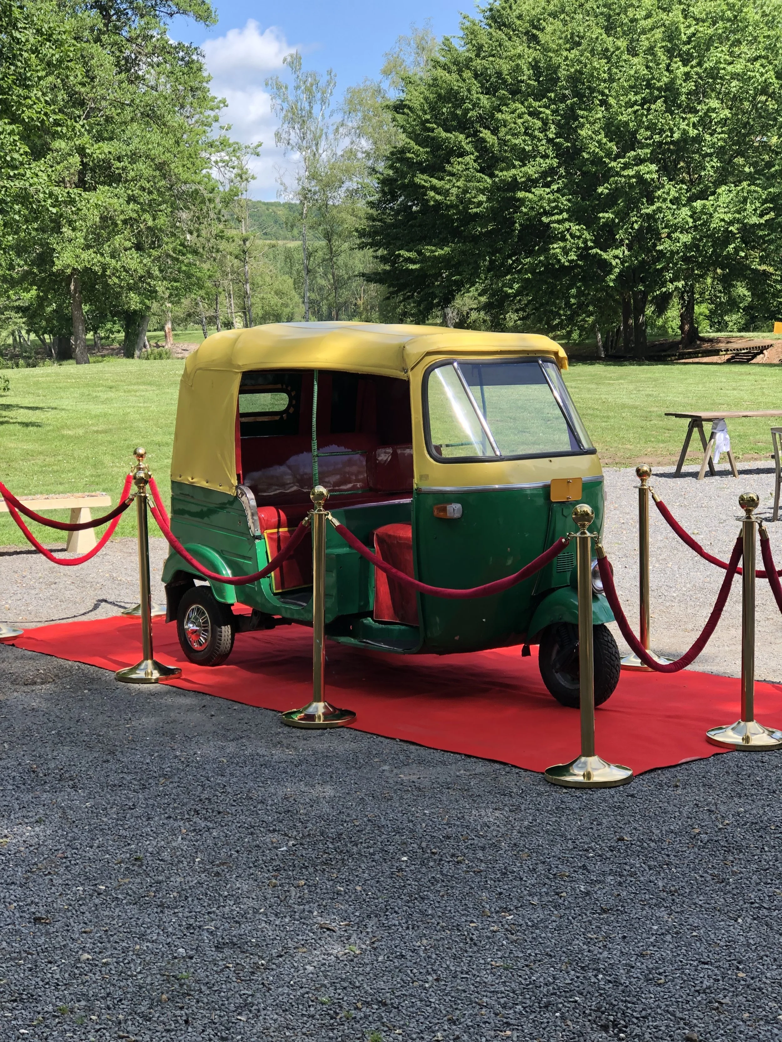 Un tuk-tuk vert et jaune garé sur un tapis rouge, entouré de barrières dorées, avec un fond de parc verdoyant.