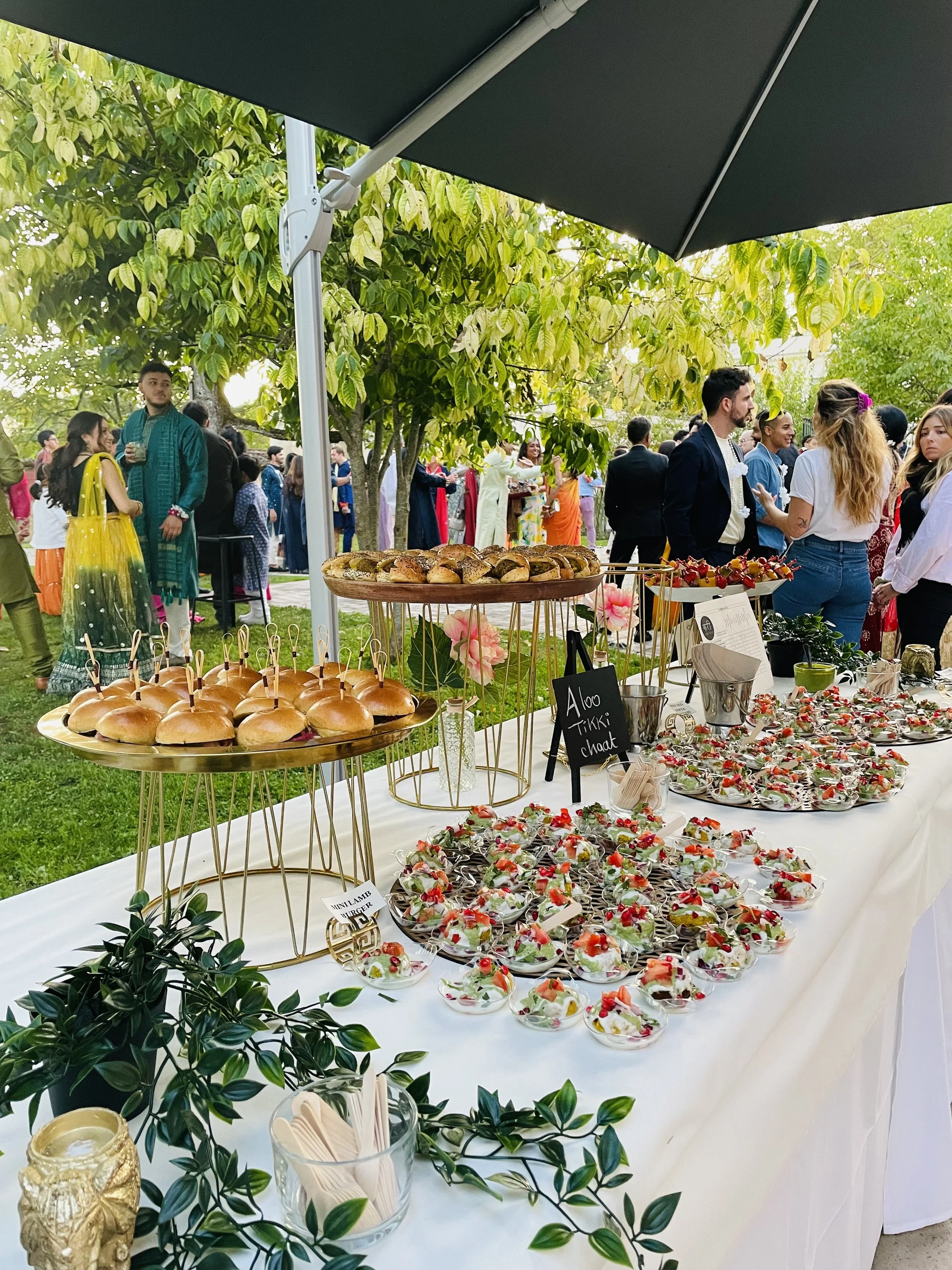 Buffet de desserts et petits sandwichs lors d'une fête en plein air avec des invités et un arbre verdoyant en arrière-plan.
