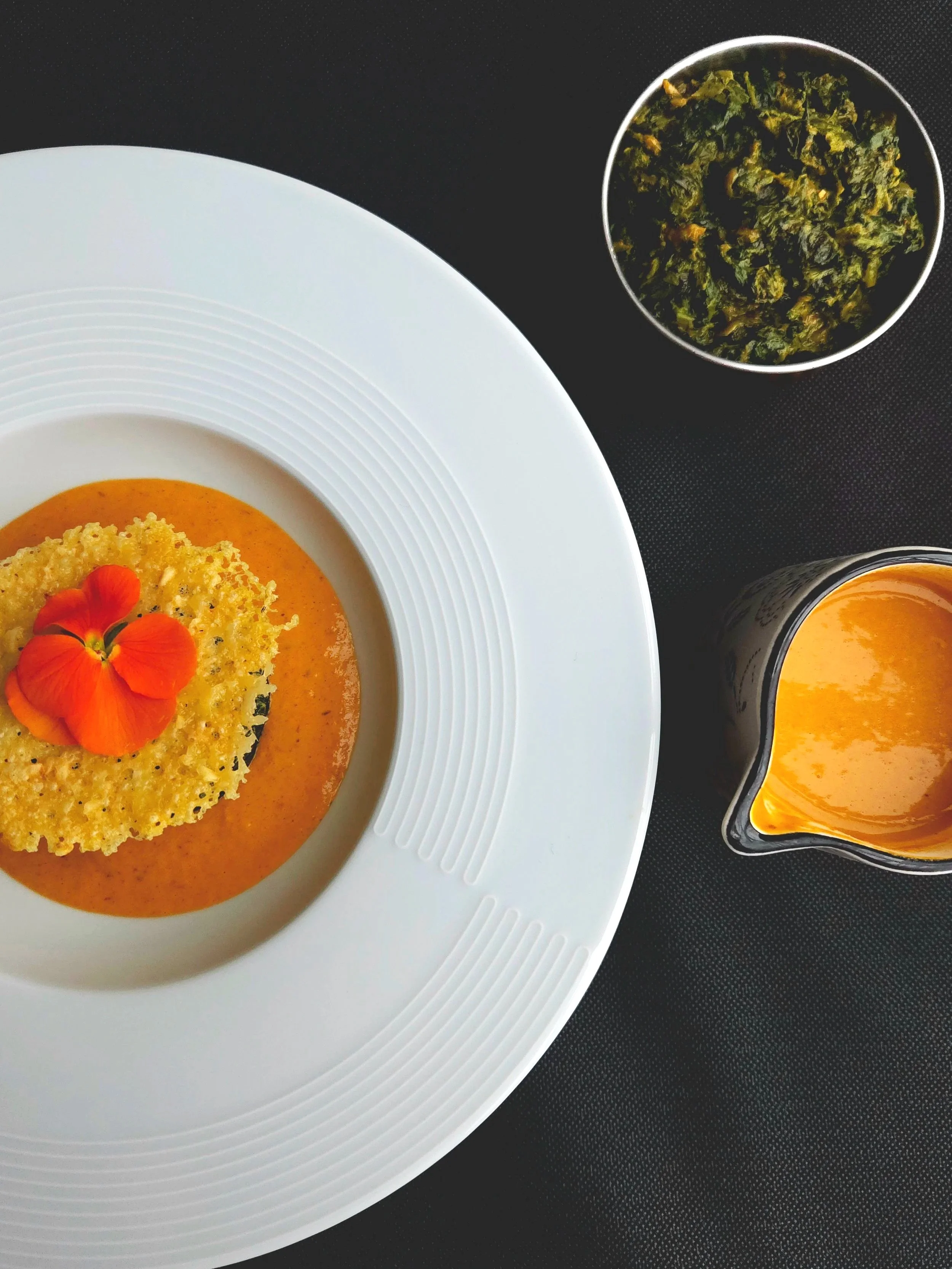 Soupe à la tomate décorée avec une tranche croustillante et des fleurs comestibles, accompagnée de feuilles de basilic et de jus d'orange.