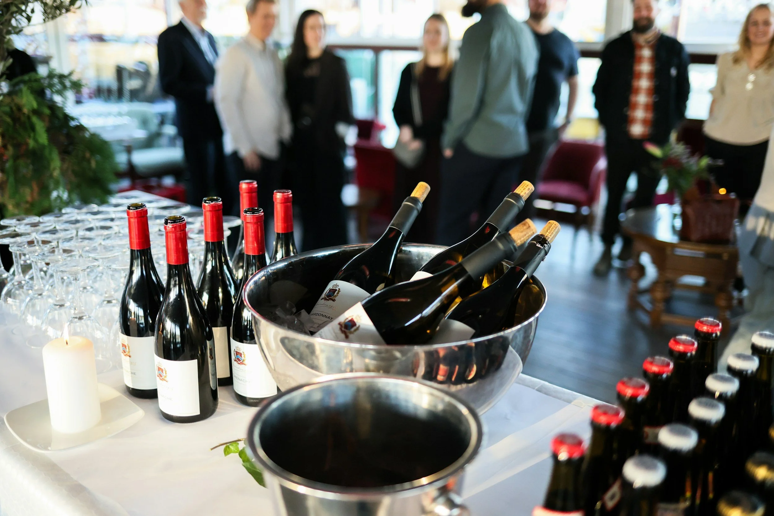 Bouteilles de vin et verres sur une table lors d'une réception, avec des personnes réunies en arrière-plan dans une salle lumineuse.