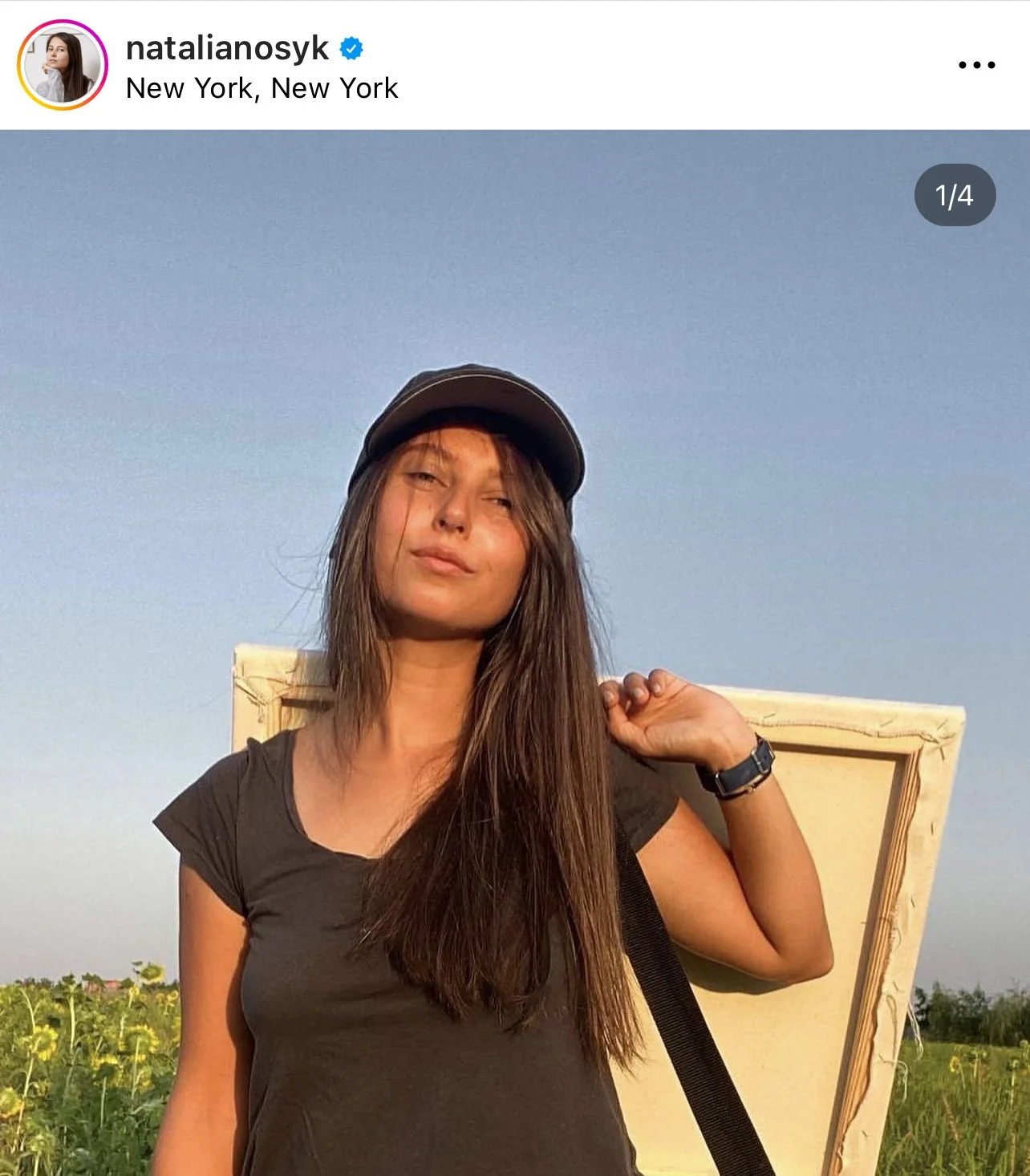 A young woman with long brown hair, wearing a black cap and black t-shirt, stands in a sunflower field at sunset. She carries an easel on her shoulder and looks into the camera with a subtle smile.