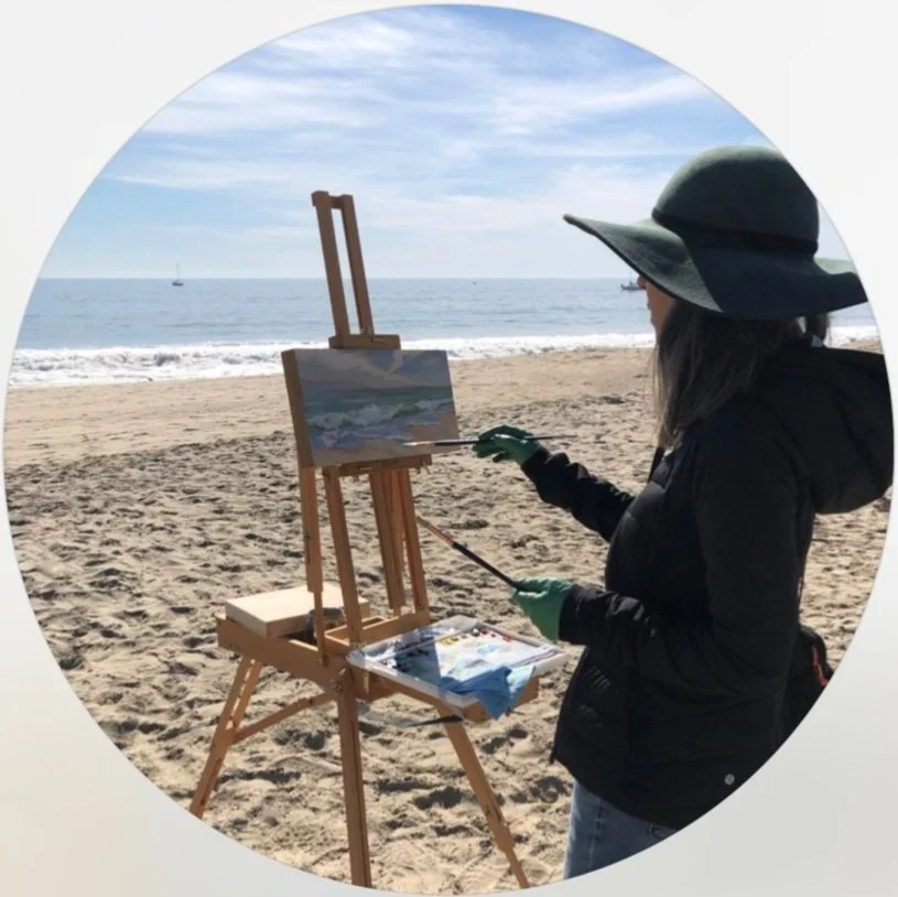 A woman painting on a canvas outdoors at the beach, wearing a wide-brim hat and gloves, with the ocean and sky in the background.