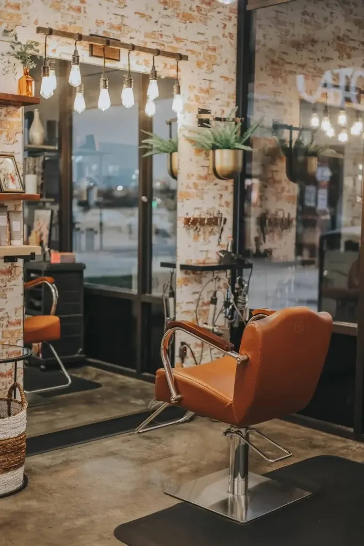 Inside a barber shop with a brown leather chair, a large mirror, hanging Edison bulb lights, and potted plants on wall-mounted planters.