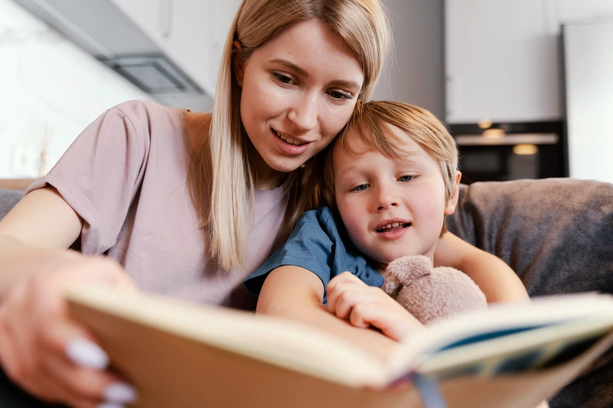 A mother sitting closely beside her young child on a couch pointing at a book during a warm daily reading practice together