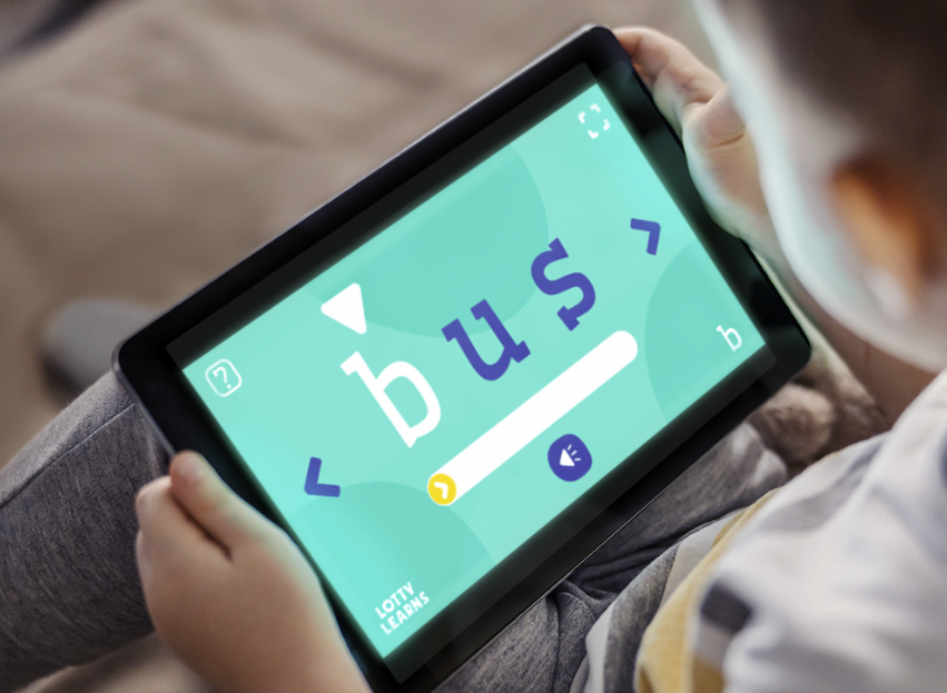 A child holding a tablet showing the Lotty Learns Phonics Factory blending tool with the word bus on screen practicing sounding out and decoding words