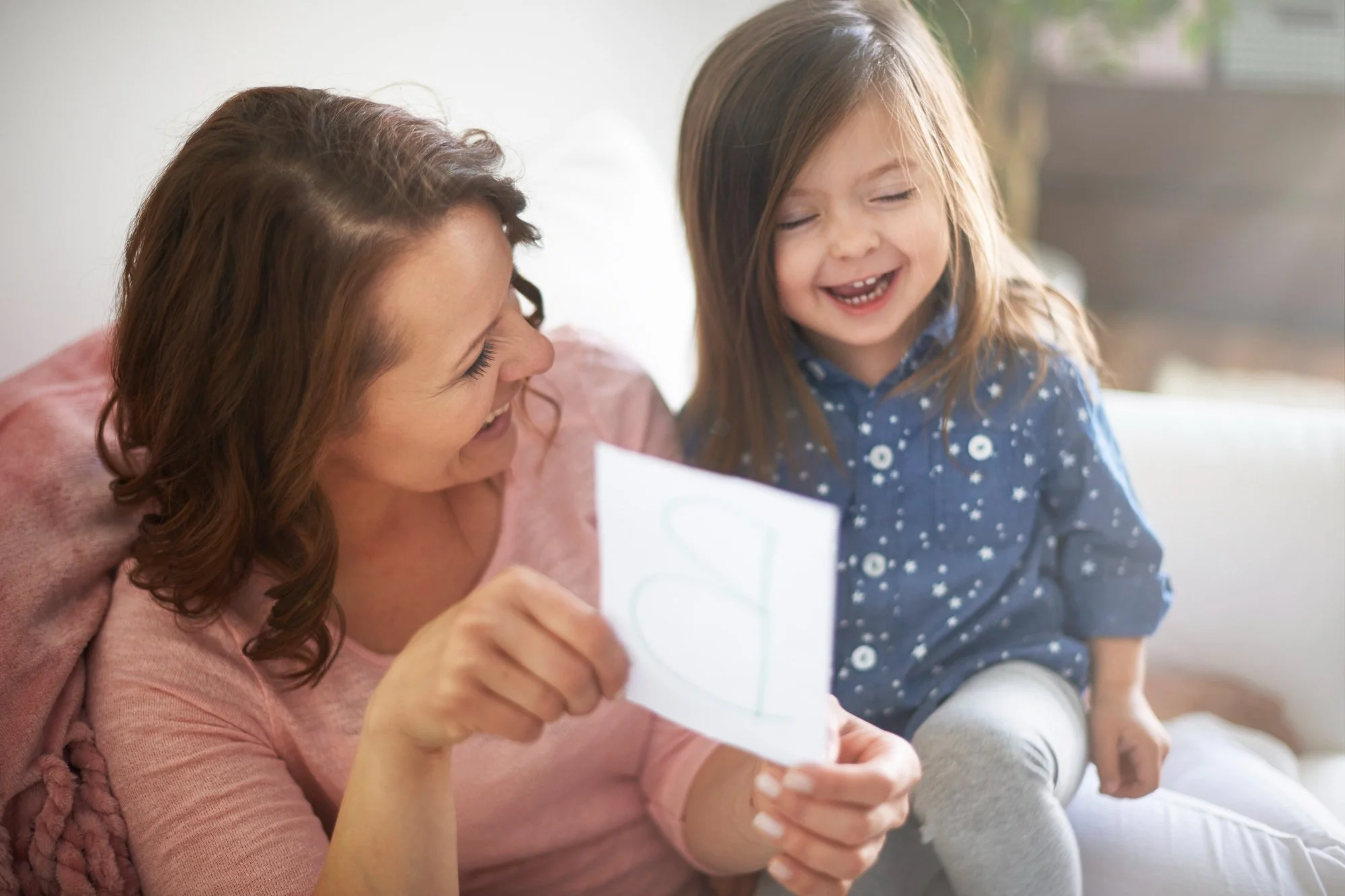 A mother holding up a piece of paper with a letter on it while her young child laughs during playful early phonics practice together