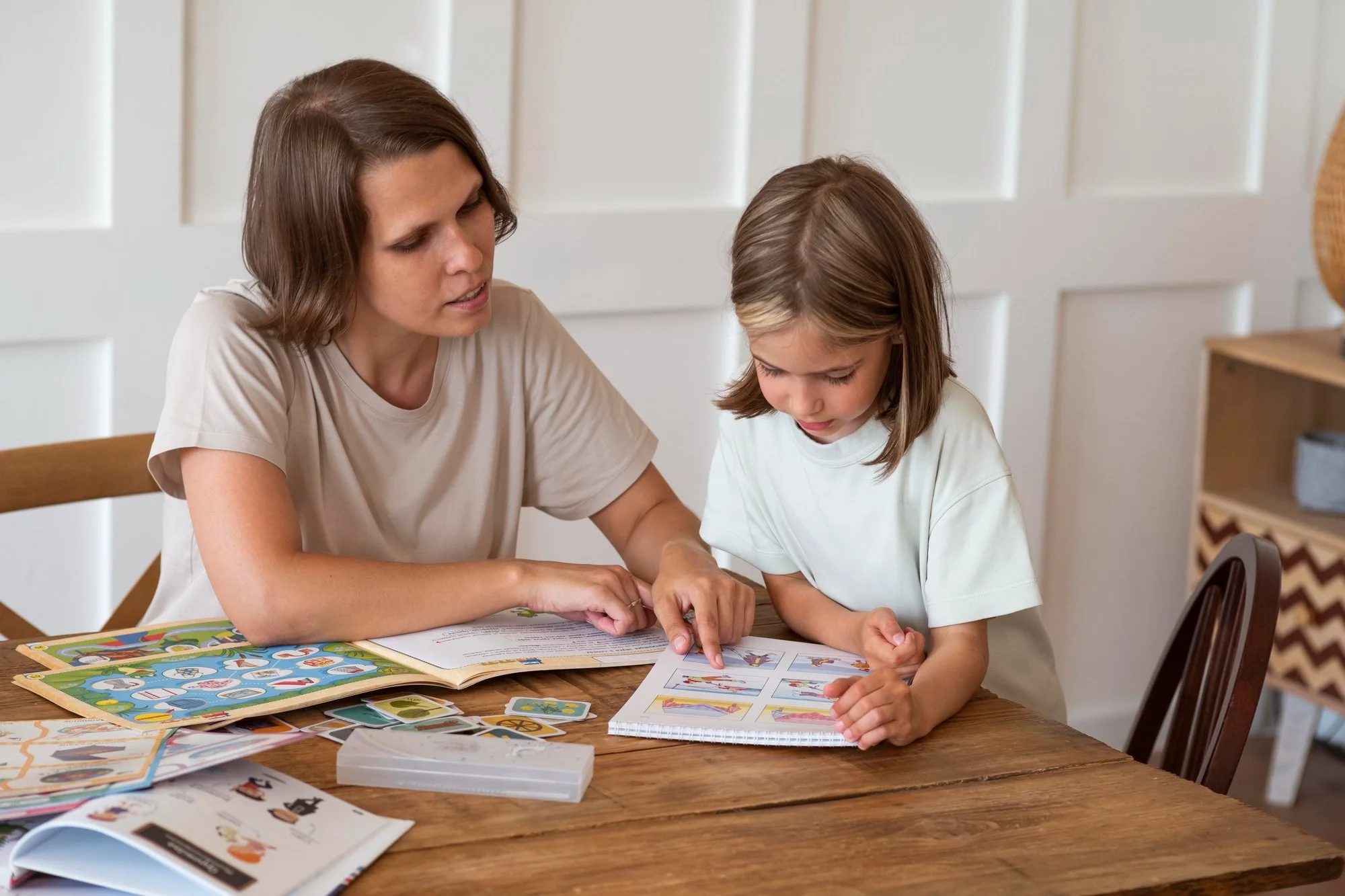 A mother sitting beside her child at a table pointing at words in a book teaching her to read and sound out words