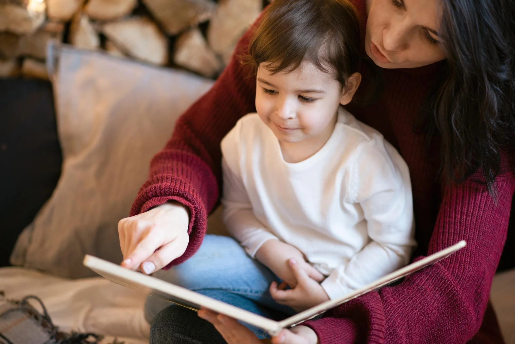 A mother sitting with her preschooler on her lap pointing at a book during early reading and phonics practice before kindergarten