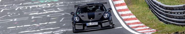 A black Porsche 911 racing car on a race track, passing through a curve with red and white striped curb and a protective barrier on the side.