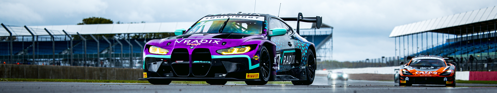 A purple and teal race car, with sponsor logos, speeding on a race track with a group of other race cars in the background.