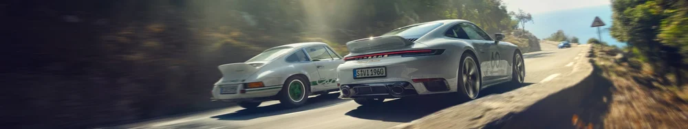 Two Porsche sports cars driving on a winding mountain road, sunlight filtering through trees.