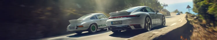 Two Porsche sports cars driving on a winding mountain road, sunlight filtering through trees.
