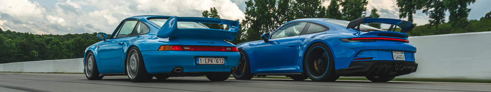 Two blue sports cars, a classic Porsche 911 Carrera RS on the left and a modern Porsche 911 Turbo on the right, racing side by side on a track with a white barrier and green trees in the background.