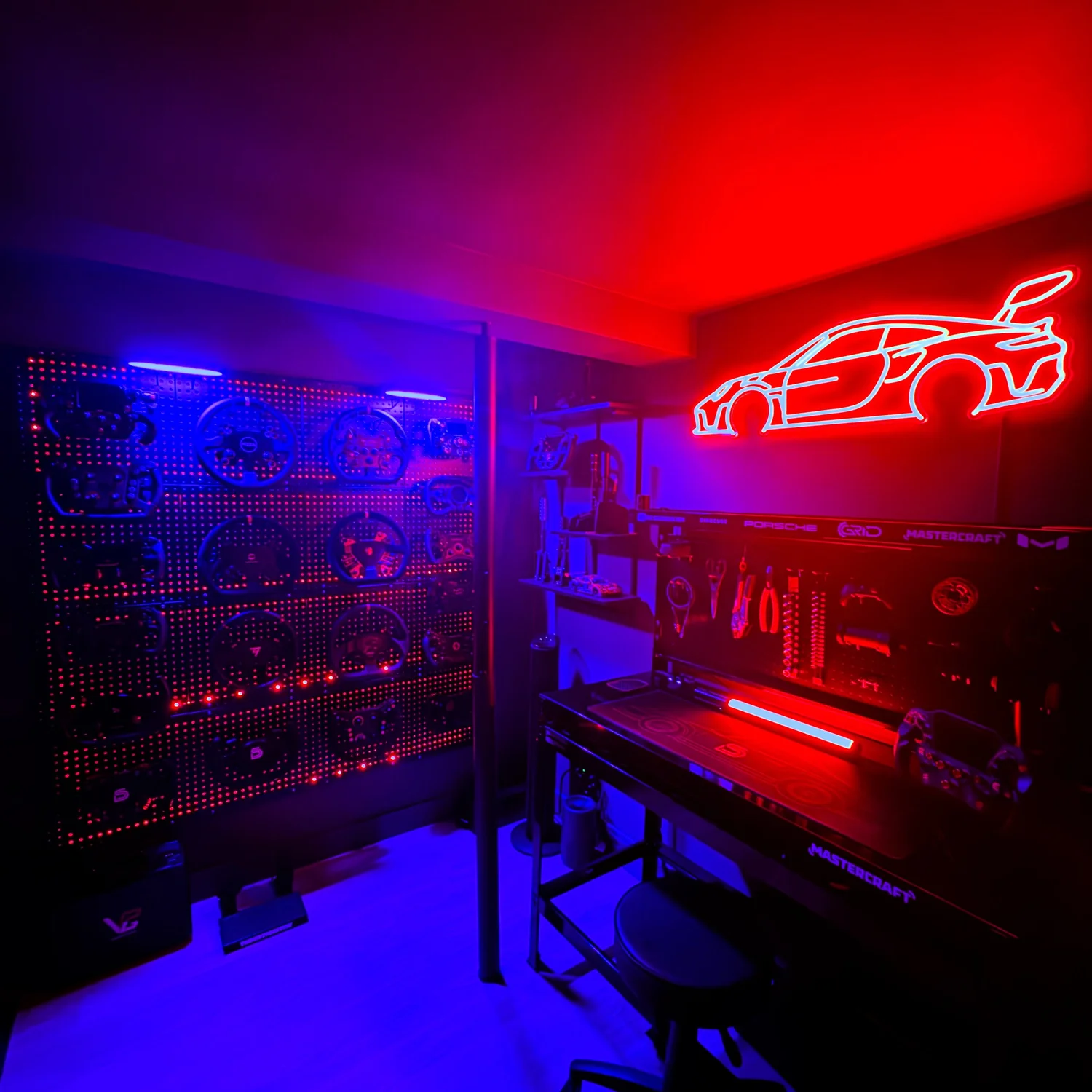 A room decorated with neon signs and automotive-themed decor. There is a red neon outline of a sports car on the wall, and a blue and red LED display of steering wheels on a pegboard. A workbench with tools and a stool are also visible, and the overall lighting is dark with vibrant neon accents.