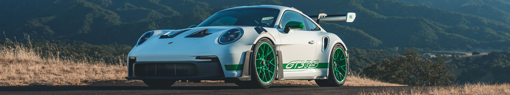 A white Porsche race car with green accents and green wheels, marked with 'GT3 RS,' is parked on a rural road with rolling hills in the background.