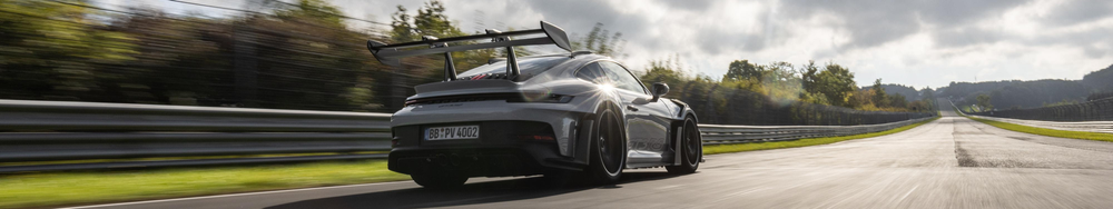 A silver Porsche race car driving on a race track with a large rear wing in a rural setting under a partly cloudy sky.