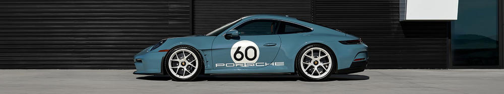 Blue Porsche sports car with racing decals, including a large white circle with the number 60 and the Porsche logo, parked on a concrete surface in front of a black building with horizontal paneling.